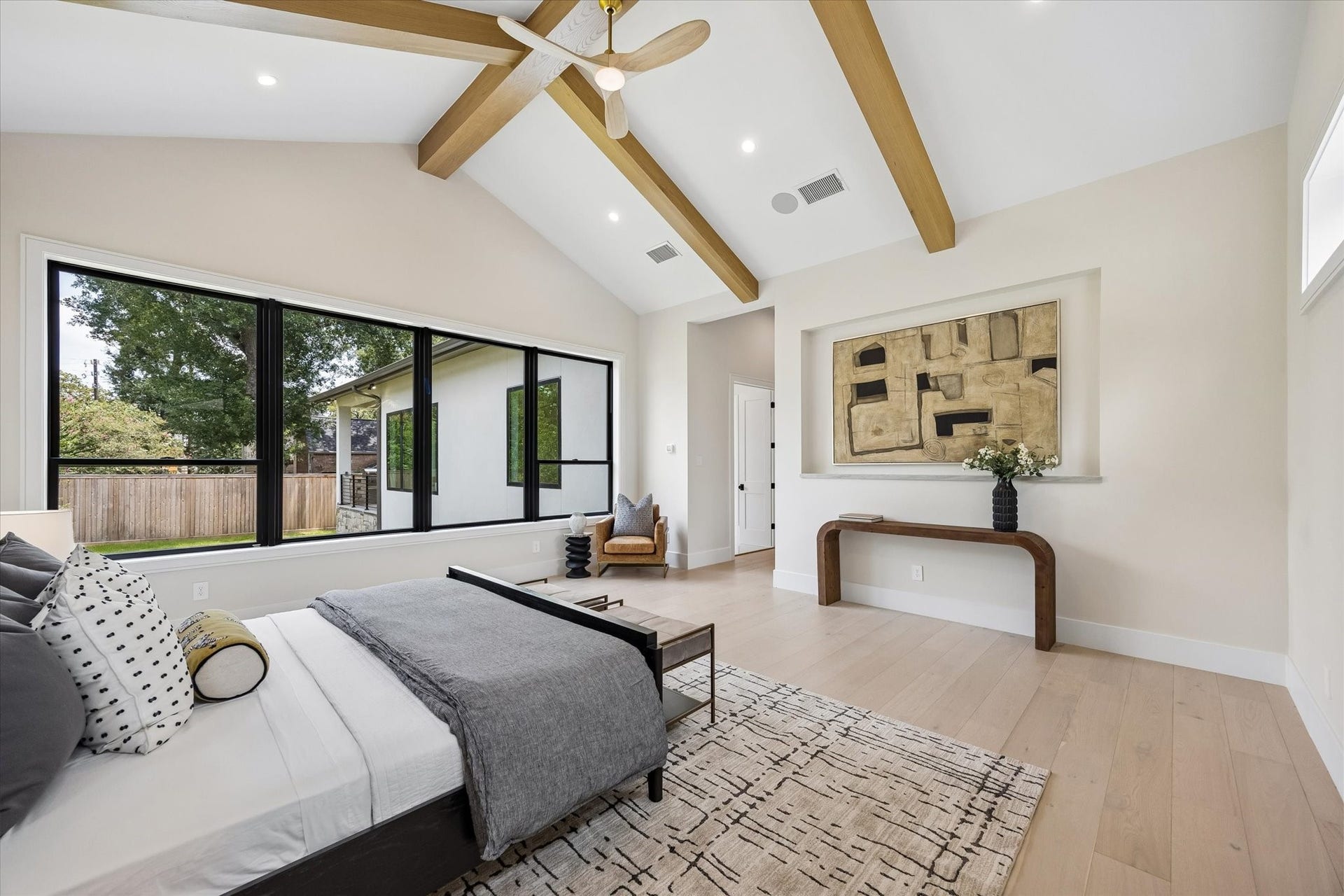 14106 Heatherfield Drive, Houston, TX 77079 The primary bedroom is a peaceful retreat defined by volume, light and understated luxury. A vaulted ceiling with warm exposed beams creates architectural interest overhead, while an expansive wall of black-framed windows brings in lush outdoor views and abundant natural light. Wide-plank flooring, soft neutral finishes and generous proportions enhance the serene atmosphere, creating an elegant space with plenty of room to relax, recharge and unwind.