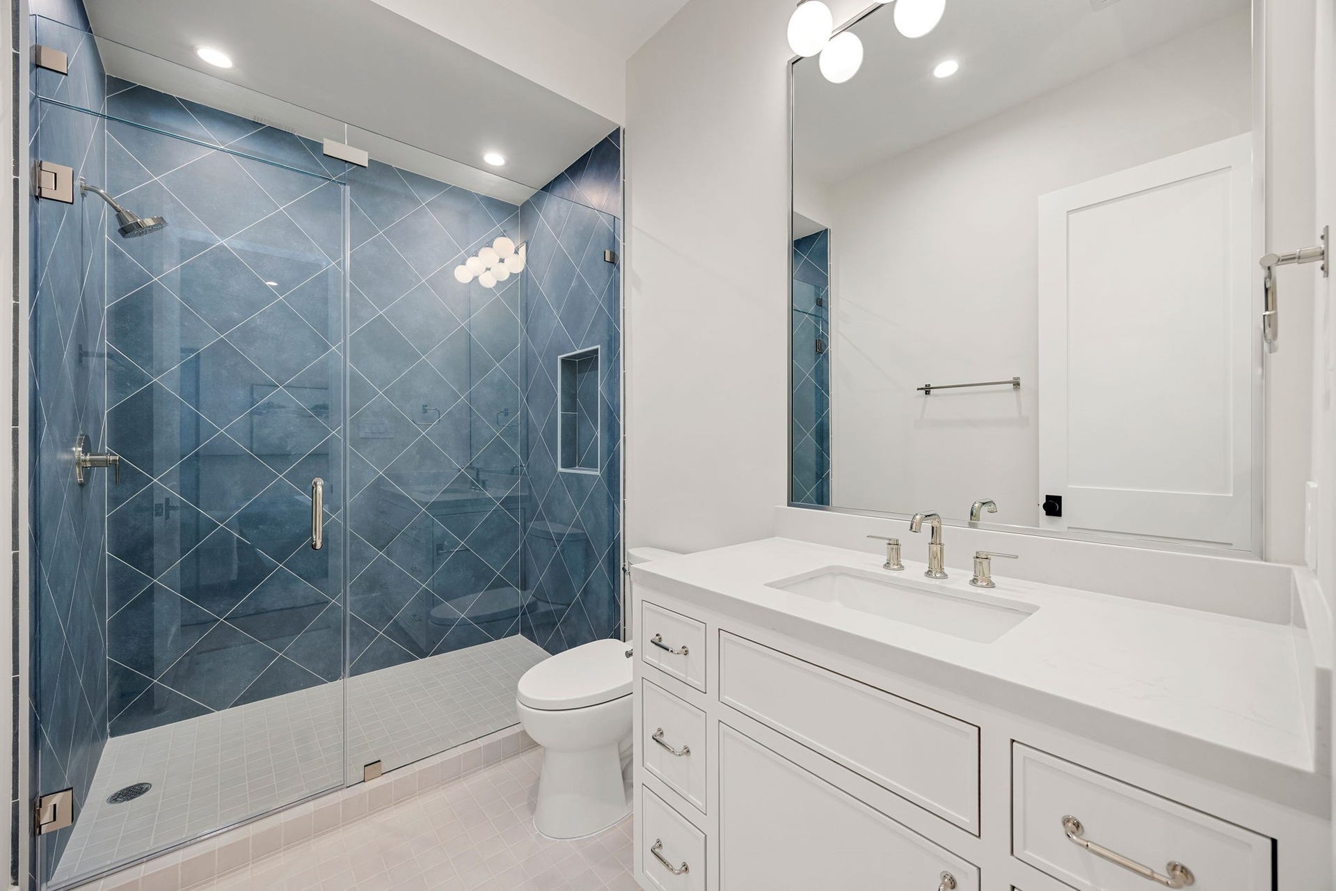 14106 Heatherfield Drive, Houston, TX 77079 A stylish and modern bathroom showcasing crisp white finishes paired with bold design elements for a striking look. The clean-lined vanity offers ample storage, while polished fixtures add a refined touch. The highlight of this space is the dramatic blue tiled walk-in shower with geometric patterning, creating a stunning focal point that elevates the entire room with contemporary flair.