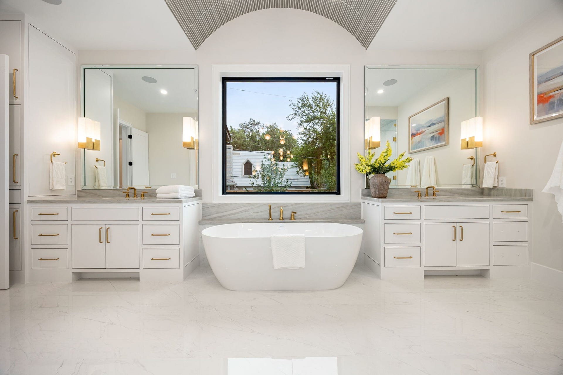 14106 Heatherfield Drive, Houston, TX 77079 A perfectly balanced view of the primary bathroom showcases symmetry and sophistication, with dual vanities flanking a central soaking tub. Large mirrors and soft lighting enhance the bright, airy feel. The highlight of this space is the centered freestanding tub beneath the window, creating a visually striking focal point that embodies both relaxation and timeless design.
