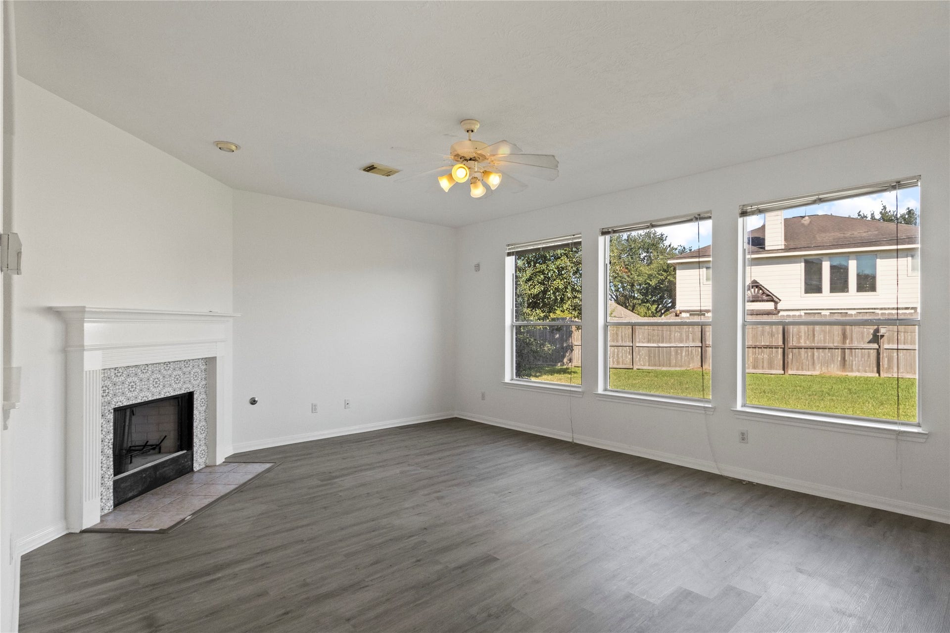 Spacious living room featuring a fireplace with decorative tile surround, gray wood-look flooring, and a ceiling fan with light fixture. Three large windows provide abundant natural light and views of the backyard. Neutral walls and an open layout offer a versatile space for relaxing and entertaining.