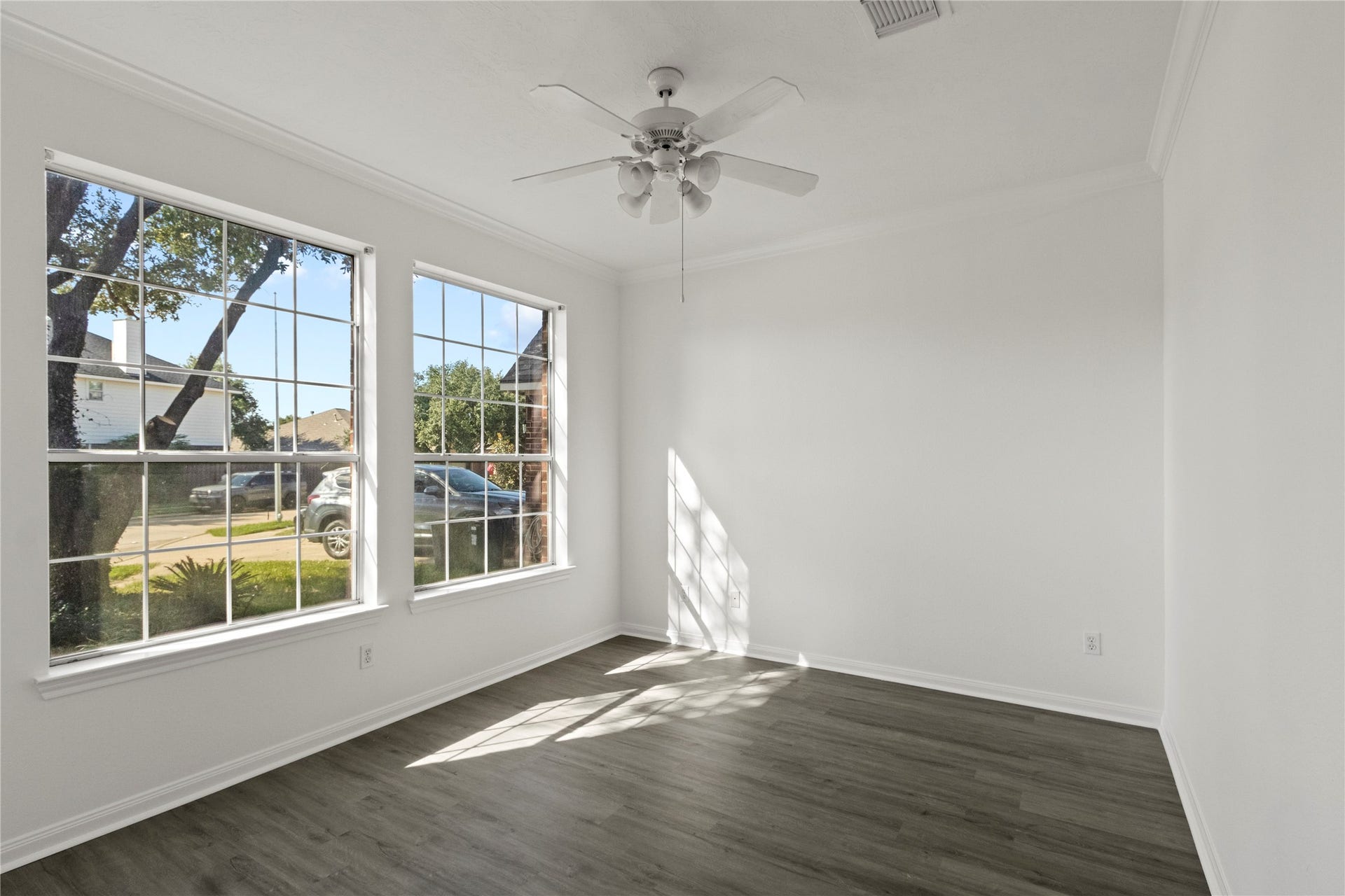 Bright home office featuring large windows that provide ample natural light, gray-toned flooring, crown molding, and a ceiling fan. The spacious layout offers flexibility for a dedicated workspace, study, or private office.