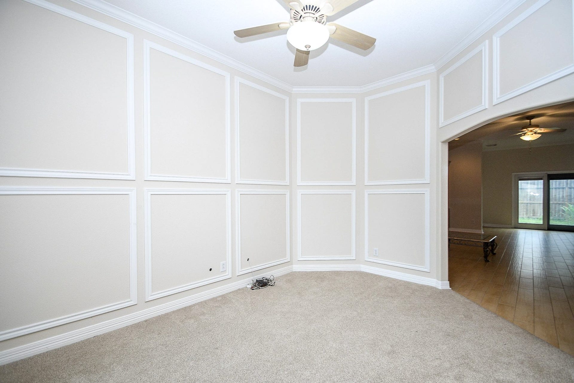 This room features neutral-toned walls with elegant paneling, a plush carpet, and a ceiling fan. It opens up to a larger space with hardwood floors and a view of the backyard through large windows, creating a bright and welcoming atmosphere.