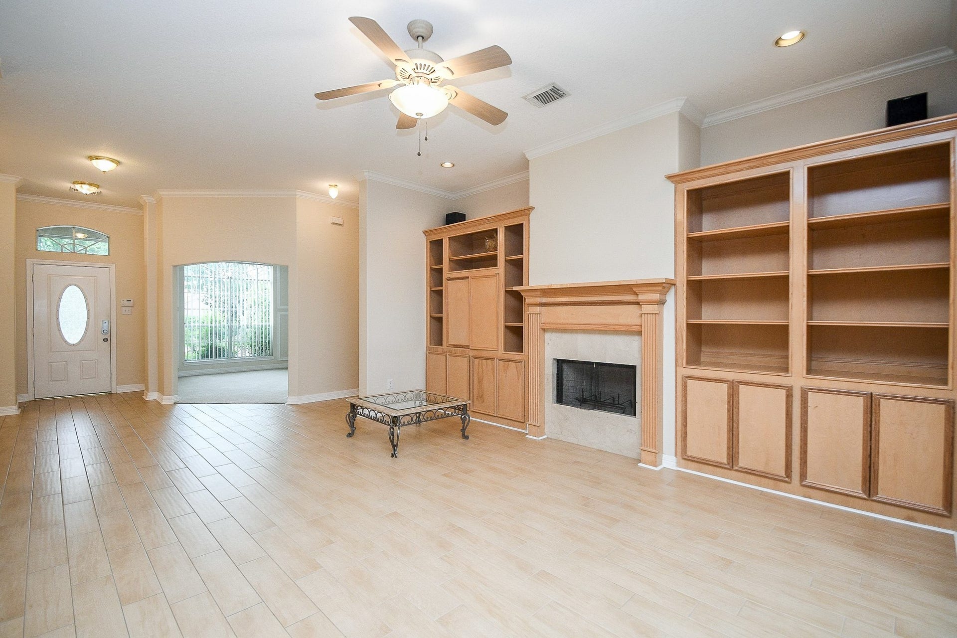 A spacious living area with light wood flooring, built-in shelves, and a fireplace. The room features a ceiling fan and recessed lighting, with a large window and an inviting front door. Ideal for cozy gatherings.