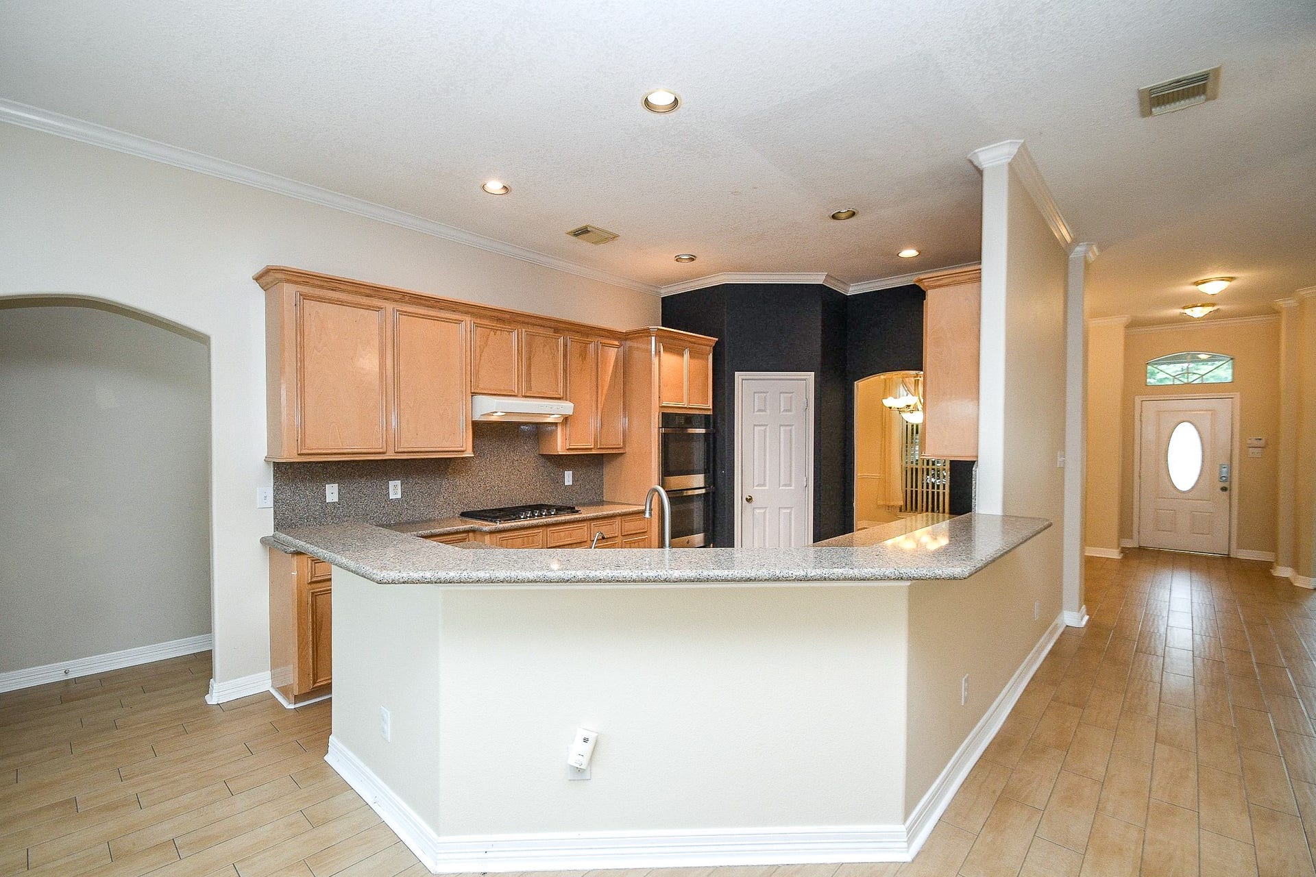 This photo showcases a spacious kitchen with light wood cabinetry, a long granite countertop, and an open layout. The kitchen leads into a bright hallway with tile flooring and a front door with a decorative window.