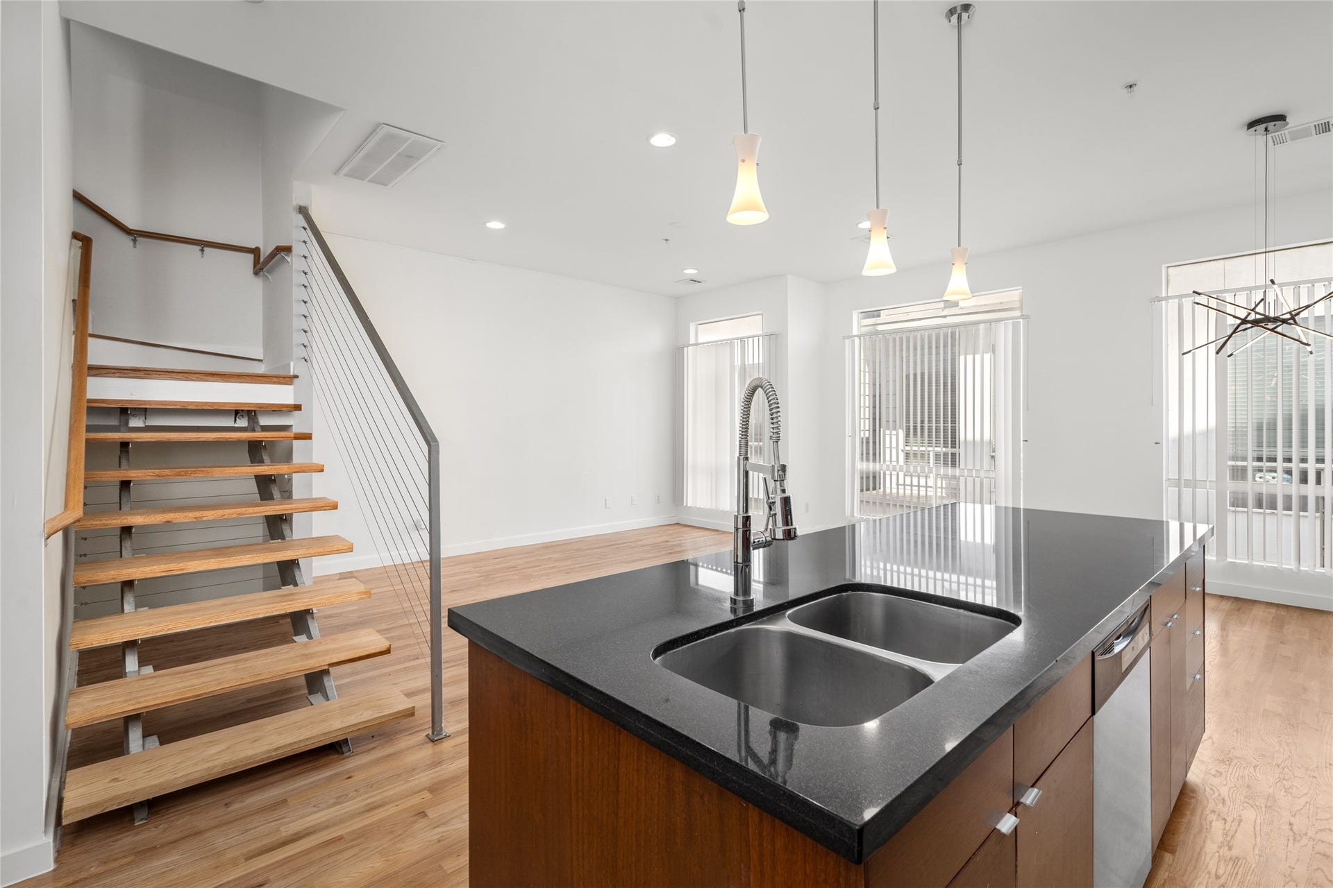 5712 Winsome Lane, #5, Houston, TX 77057 A light hardwood floor covers the space. A wooden staircase with metal railings leads up, its open design showing the support structure.
In the center, a modern kitchen island stands with a dark granite countertop, a stainless steel double sink, and a sleek faucet. The island has medium-brown wood cabinets, and a dishwasher is partly visible on the right.