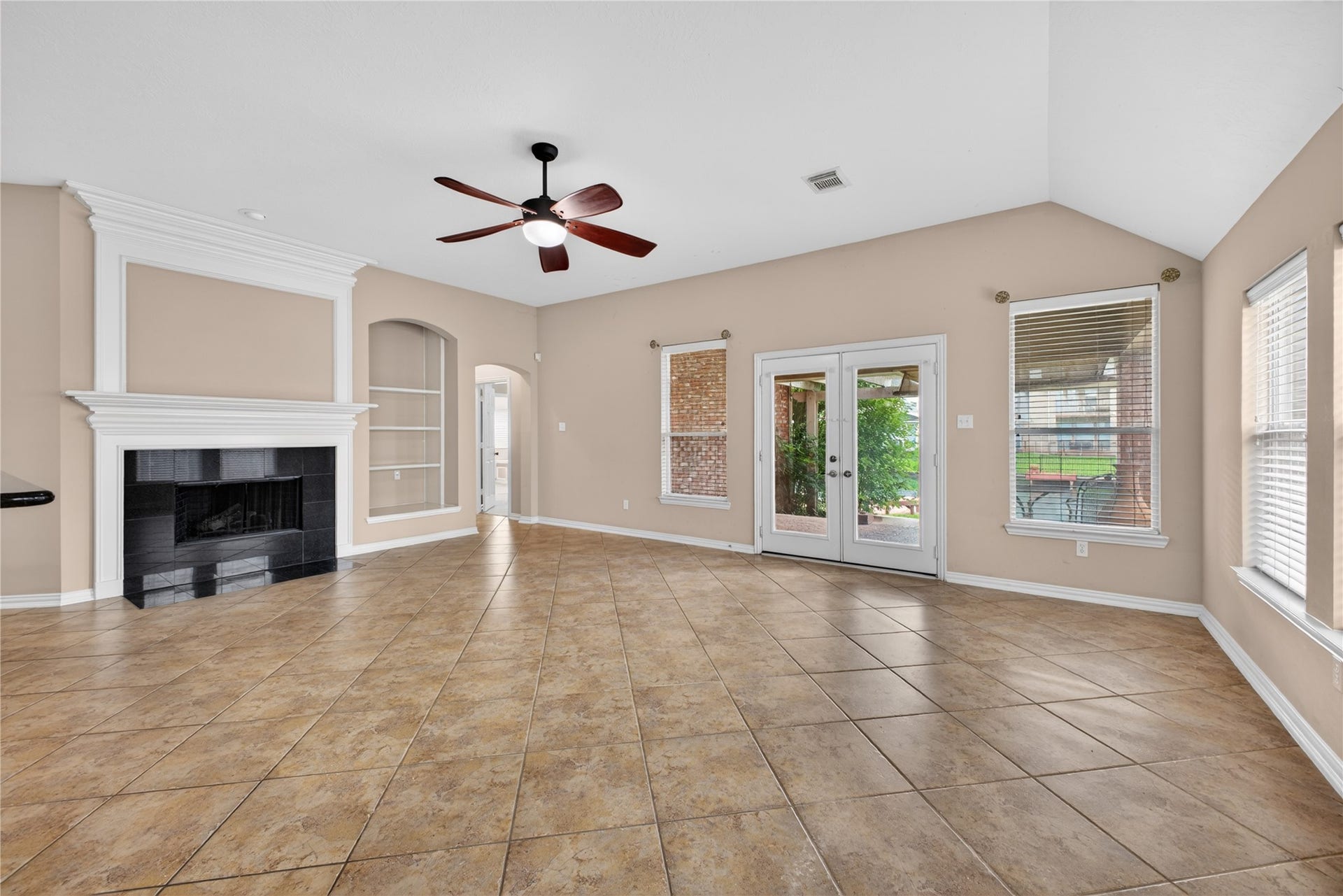 14326 Manorbier Lane, Sugar Land, TX 77498 This spacious living room features a tiled floor, neutral walls, and ample natural light from multiple windows. It includes a cozy fireplace with a white mantel, built-in shelving, and French doors leading to an outdoor area. A ceiling fan adds comfort and style.