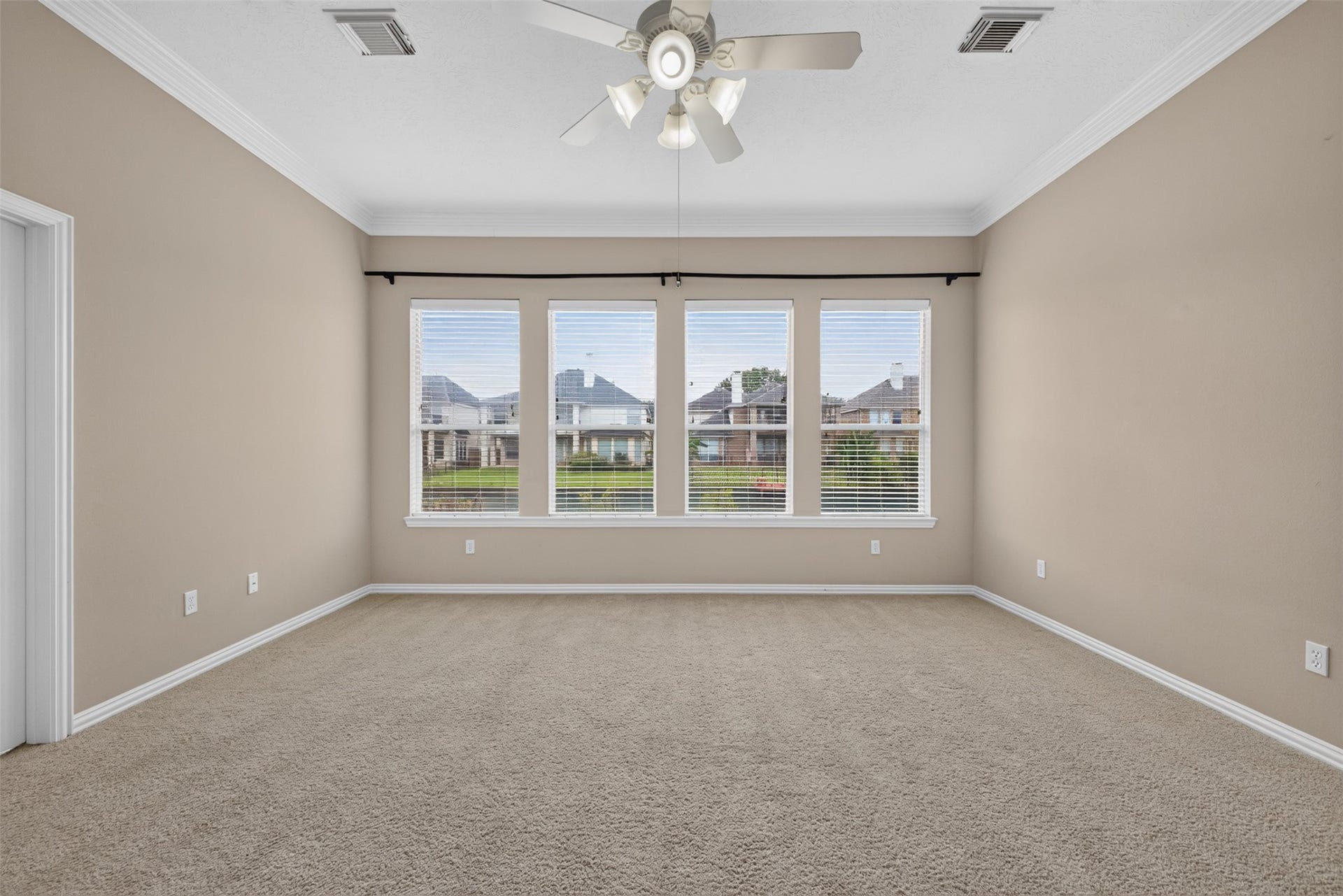 14326 Manorbier Lane, Sugar Land, TX 77498 Spacious bedroom with beige walls and carpet, featuring large windows that offer plenty of natural light. A white ceiling fan is centrally positioned, and there's crown molding for an elegant touch. Adjacent access to a bathroom is visible.