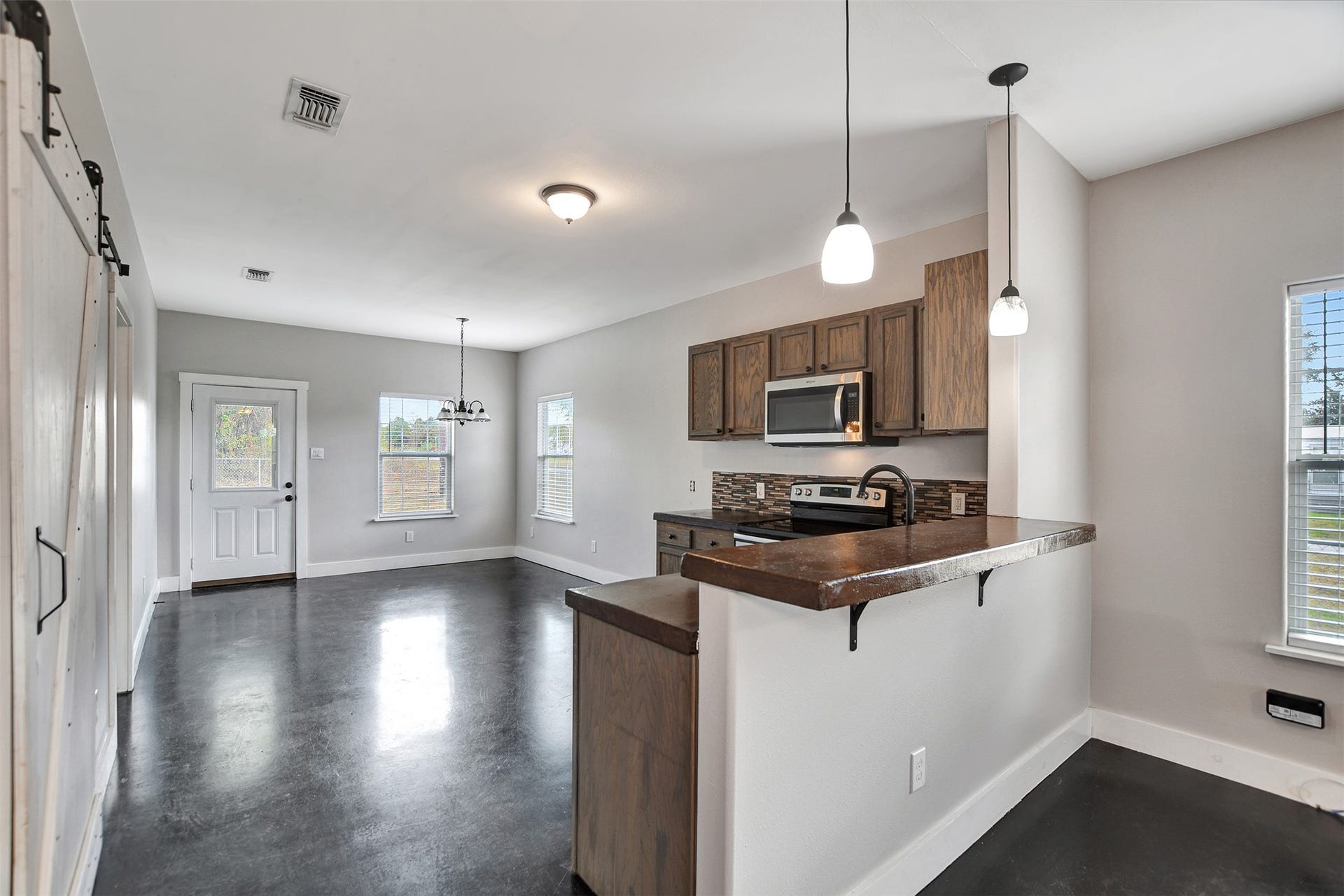 2546 16th Street, Hempstead, TX 77445 This is a modern, open-concept kitchen with wood cabinetry, stainless steel appliances, and a breakfast bar. The space is equipped with pendant lighting and has ample natural light coming through multiple windows, along with sleek, polished concrete floors.