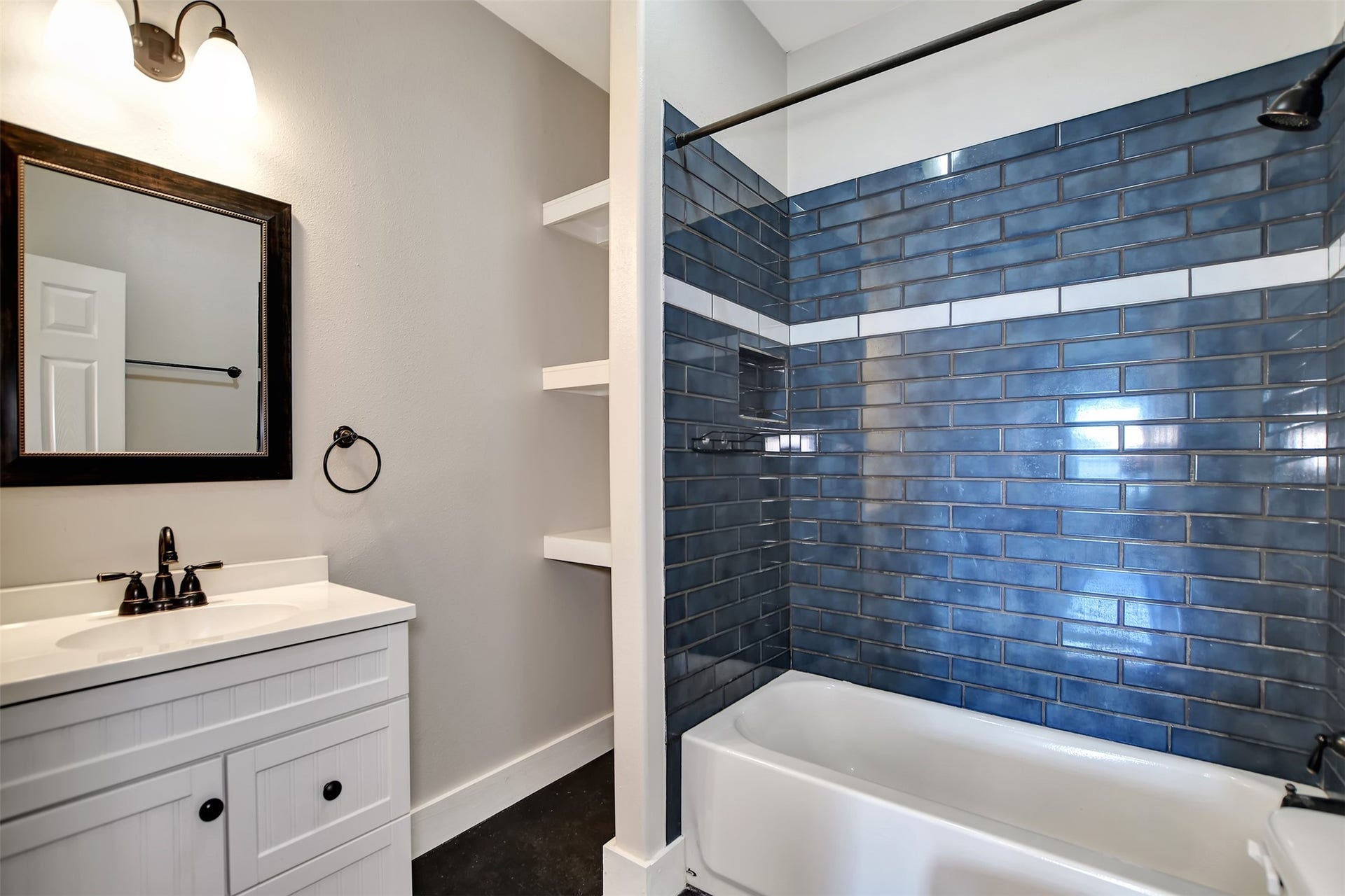 2546 16th Street, Hempstead, TX 77445 Modern bathroom featuring a white vanity with a dark countertop, a large mirror, stylish navy blue subway tiles in the shower area, built-in shelving for storage, and elegant dark fixtures.