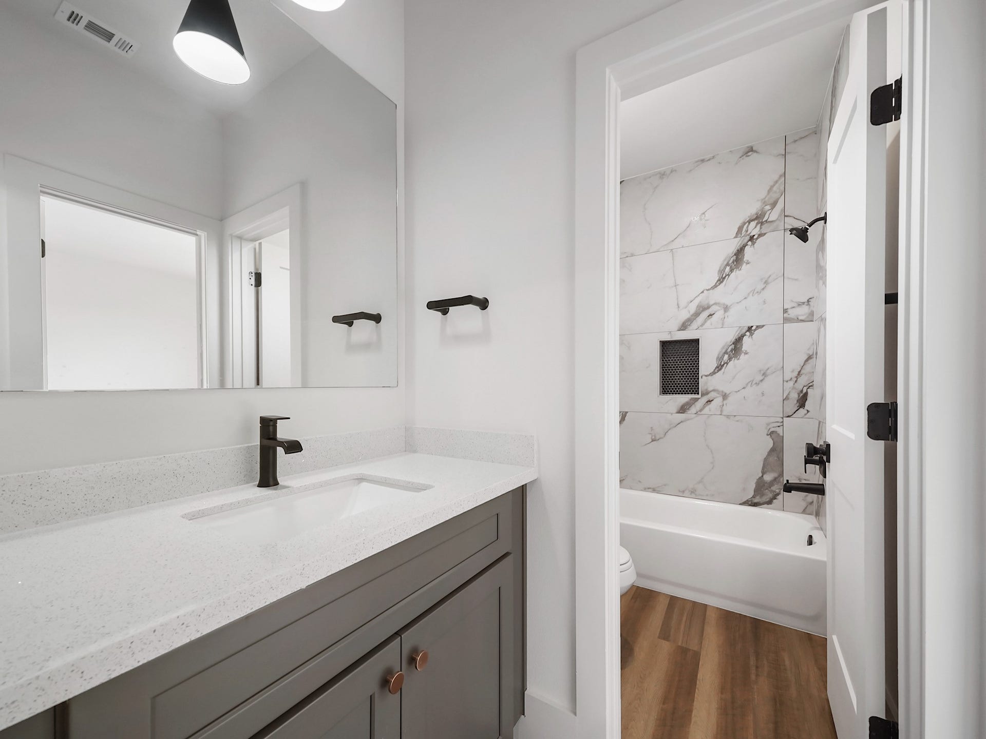 881 Rachel Street, #A, Houston, TX 77091 This modern bathroom features a sleek design with a large mirror, a quartz countertop, a stylish dark faucet, and ample under-sink storage. The adjacent shower area has elegant marble-style tiles and a convenient built-in shelf. Wood-look flooring adds warmth to the space.
Photo Disclaimer: Photos are representative of the community and may depict a similar floor plan, model home, or previously completed residence.