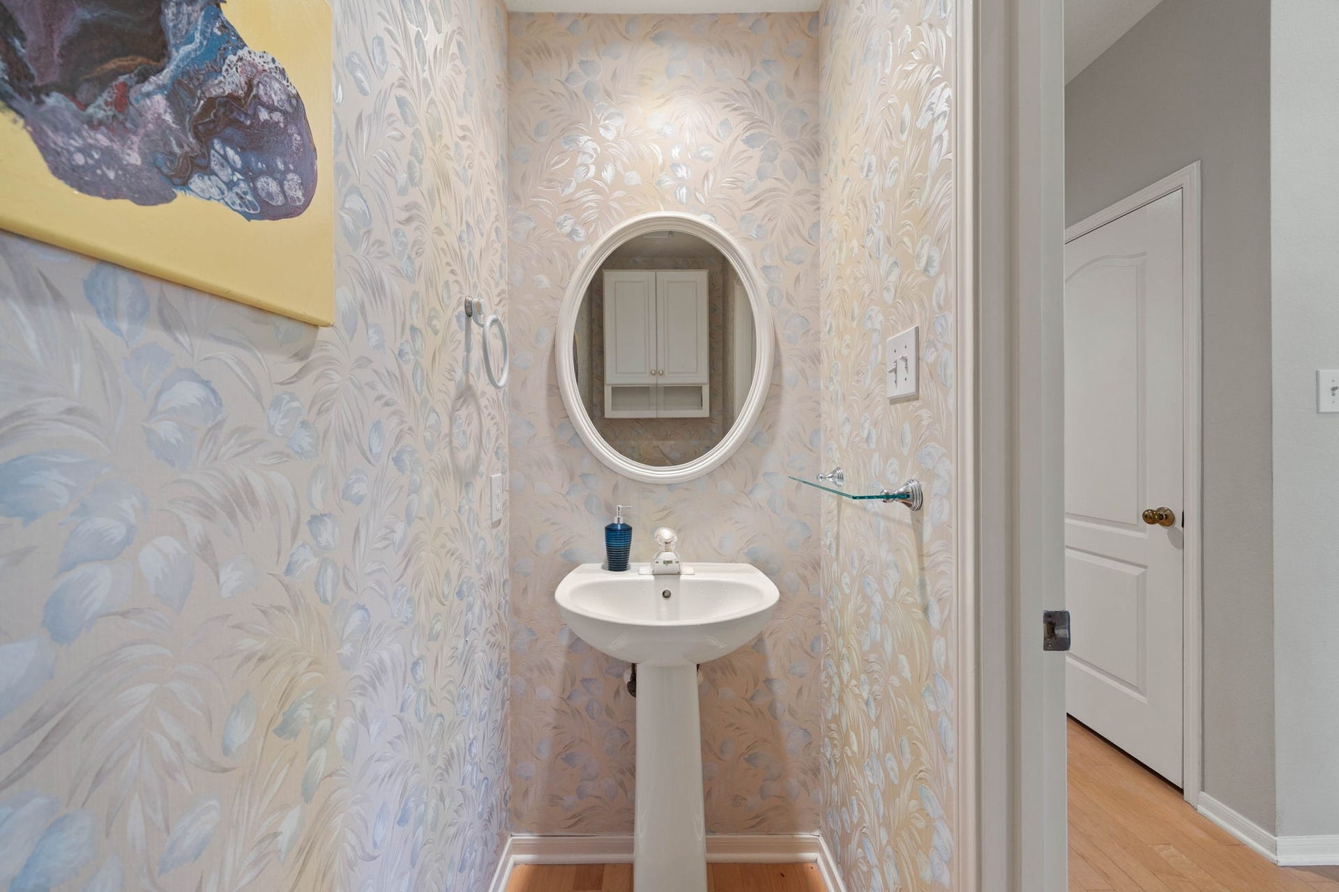 205 Purple Slate Place, The Woodlands, TX 77381 This charming first-floor powder room makes a stylish statement, wrapped in elegant, patterned designer wallpaper. Perfect for guests, the space is beautifully optimized with a classic white pedestal sink, a matching white-framed oval mirror, and a sleek glass utility shelf.