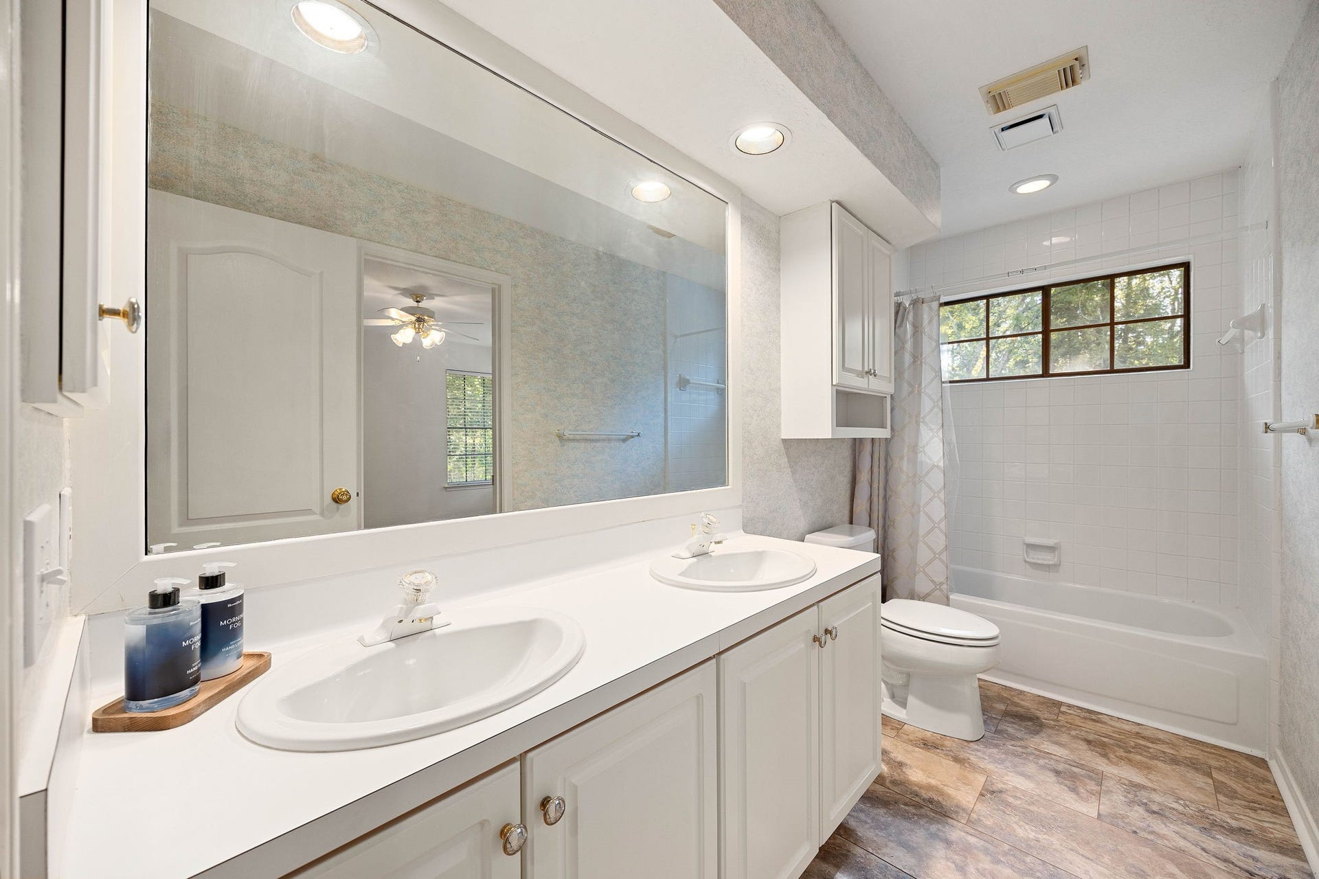 205 Purple Slate Place, The Woodlands, TX 77381 The secondary en-suite bathroom is shared with a secondary bedroom and game room areas. It showcases a spacious double-sink vanity with crisp white cabinetry, mirror, recessed and soffit lighting, while a separate built-in medicine cabinet and upper wall cabinet provide generous storage. A classic tub/shower with tile surround features a bright window that pulls in natural light.