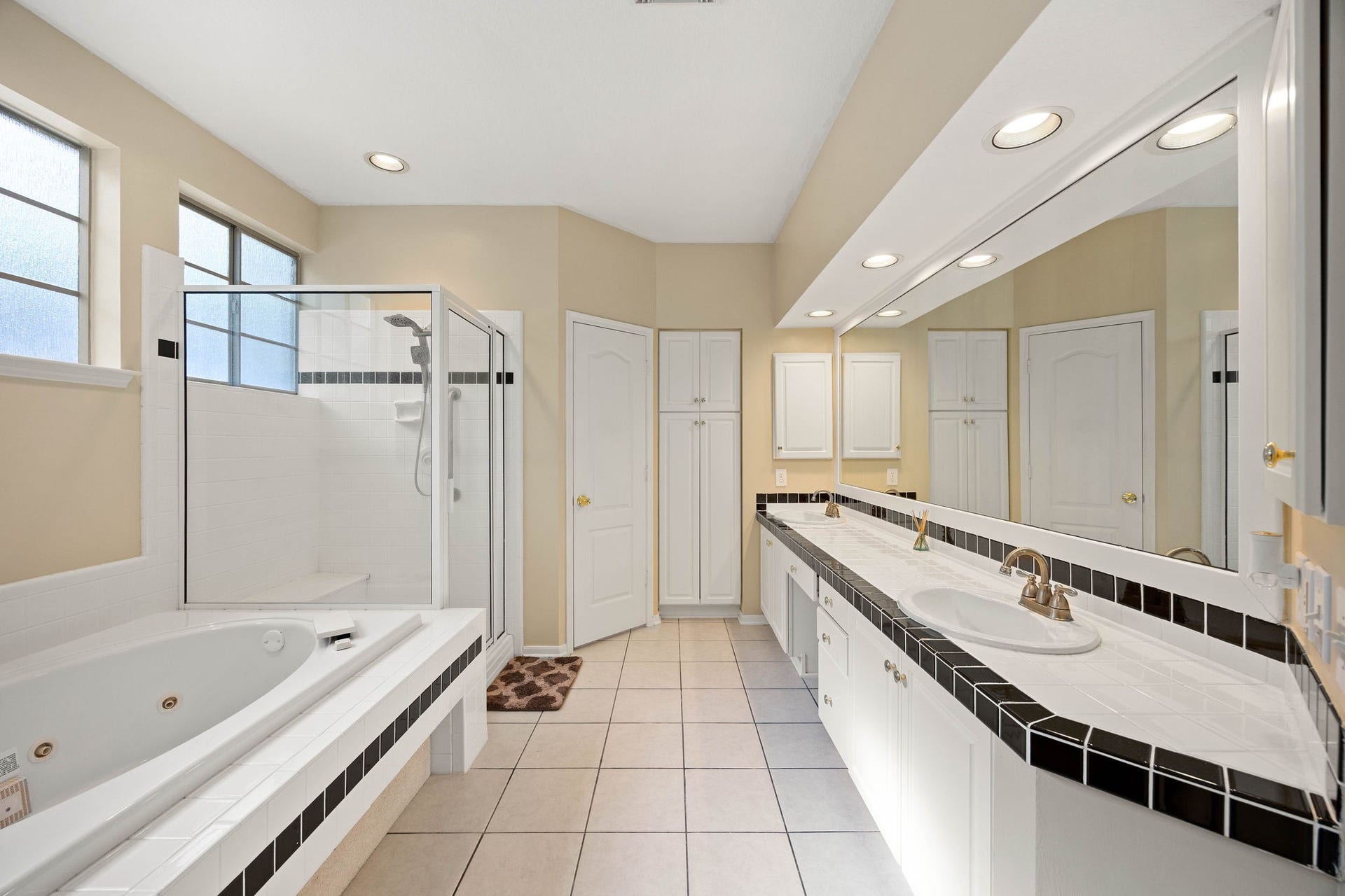 205 Purple Slate Place, The Woodlands, TX 77381 The spacious primary bathroom serves as a spa-like retreat, showcasing an oversized jetted soaking tub and a separate glass-enclosed shower. A massive dual-sink vanity features crisp white cabinetry, a built-in makeup station, and a wide mirrored wall illuminated by integrated soffit lighting. The room is detailed with a classic black-and-white tile trim, durable ceramic floor tile, a frosted glass window for privacy, and a custom built-in linen tower paired with a toilet compartment.
