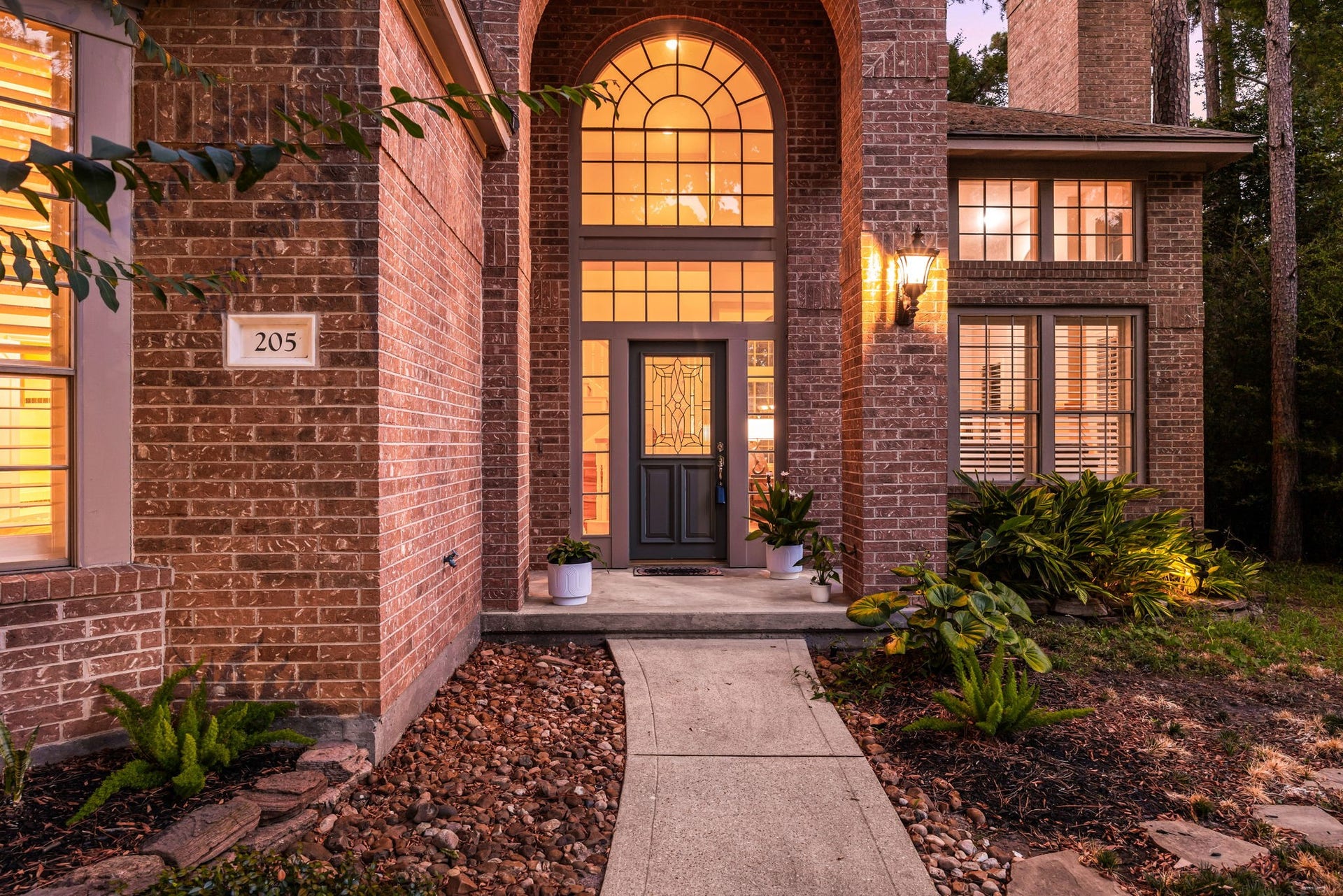 205 Purple Slate Place, The Woodlands, TX 77381 Step up the concrete walkway and into a grand entrance designed to impress. The soaring brick archway frames a handsome decorative front door, while multi-pane upper windows cast a welcoming glow across the front estate. Thoughtful details like the classic carriage lantern and lush perennial landscaping establish a sophisticated yet deeply welcoming atmosphere before you even step inside.