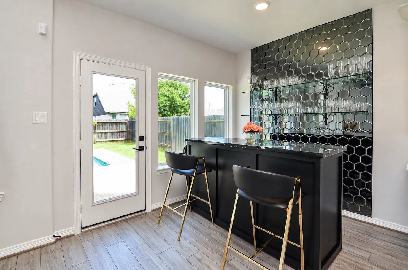 30706 Hawthorn Glen Court, Fulshear, TX 77441 Stylish home bar with sleek black hexagonal backsplash, well-stocked glass shelves, and a chic counter overlooking the backyard pool area. Ideal for entertaining.