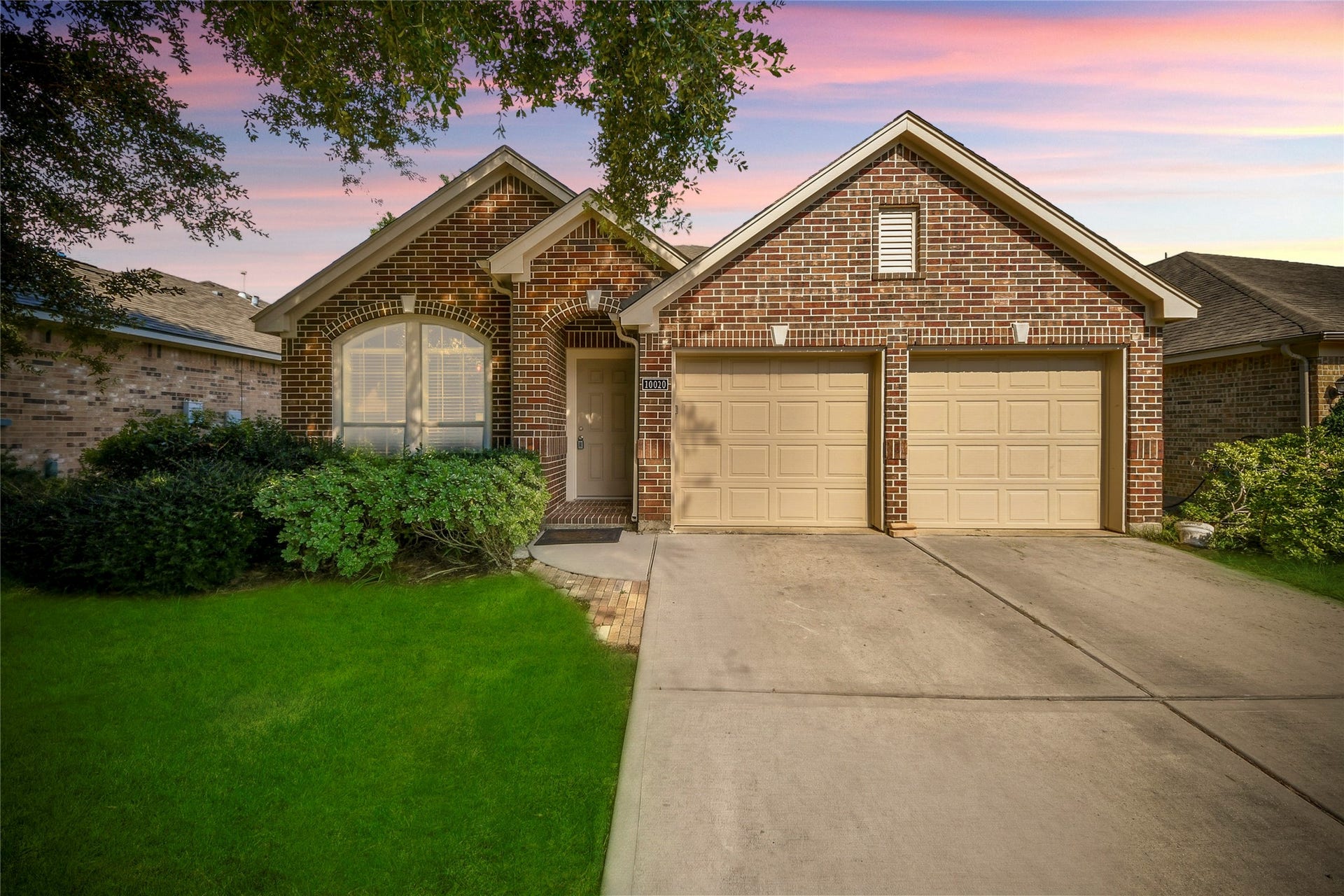 Charming brick home with a welcoming arched entryway and a spacious two-car garage, set against a lush green lawn and a serene sky.