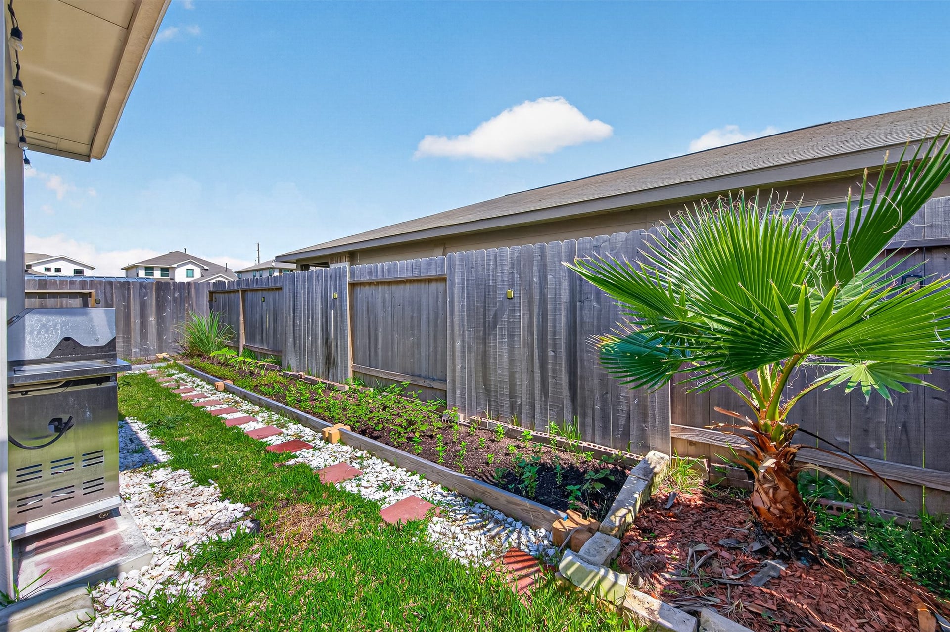 22409 Kinley Street, New Caney, TX 77357 Charming backyard with a wooden fence, featuring a small garden bed and a palm plant. Includes a BBQ grill area, perfect for outdoor cooking.