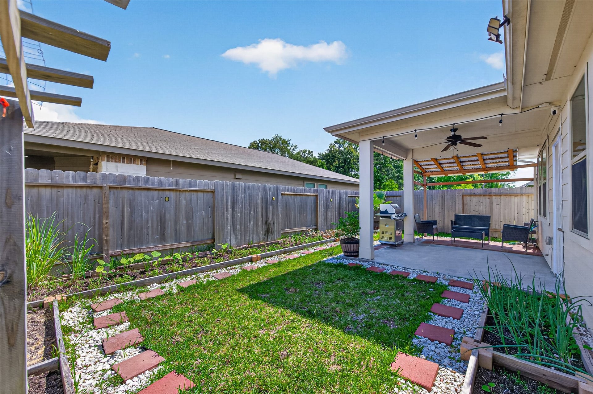 22409 Kinley Street, New Caney, TX 77357 This backyard features a cozy covered patio with seating and a ceiling fan, ideal for outdoor relaxation. A well-maintained garden borders the grassy area, and stepping stones lead across the yard. The space is enclosed by a wooden fence for added privacy.