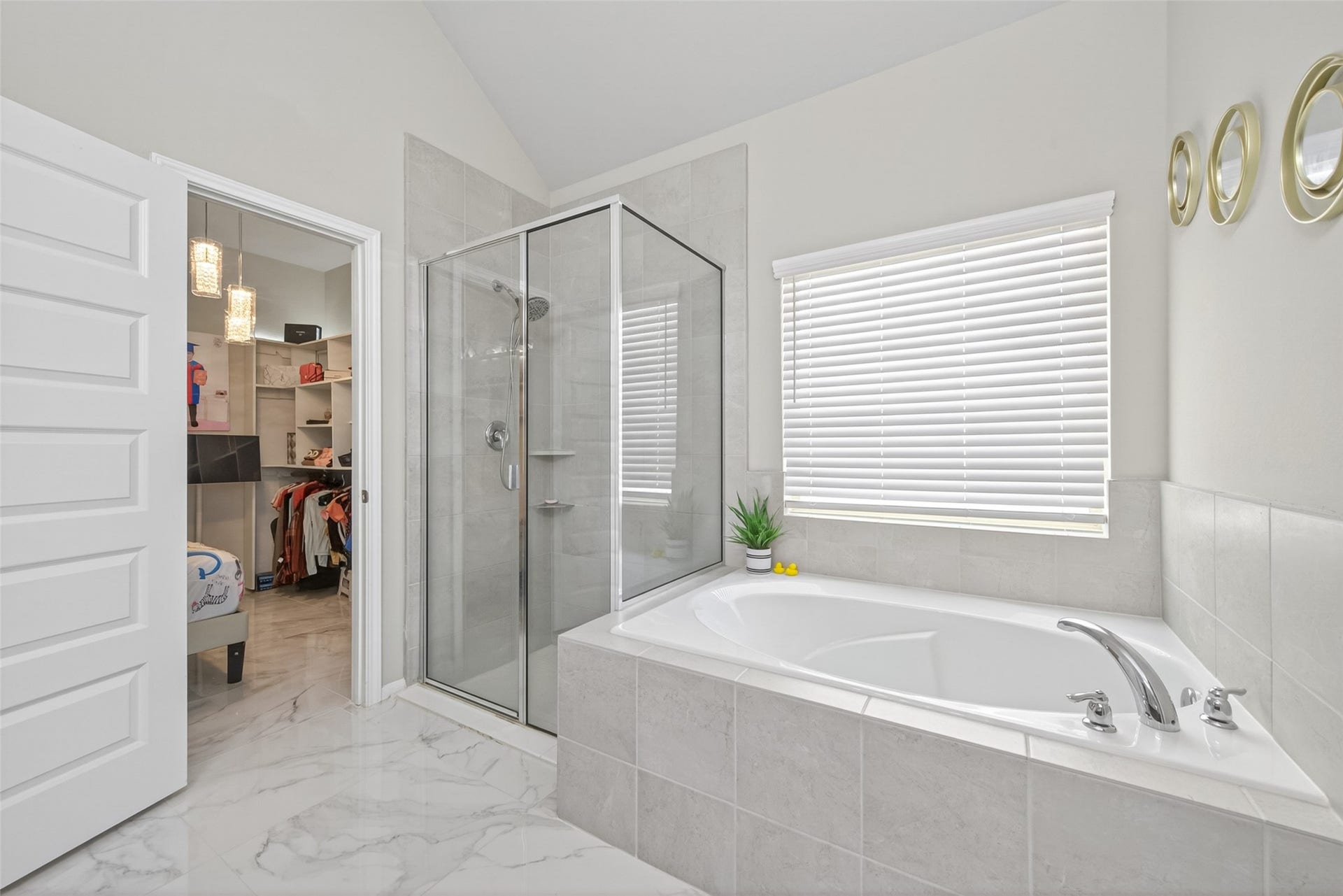 22409 Kinley Street, New Caney, TX 77357 This bathroom features a modern design with a spacious corner tub, a glass-enclosed shower, and large tiles. It includes a window with blinds for natural light and privacy, and has an adjacent walk-in closet with ample storage space.