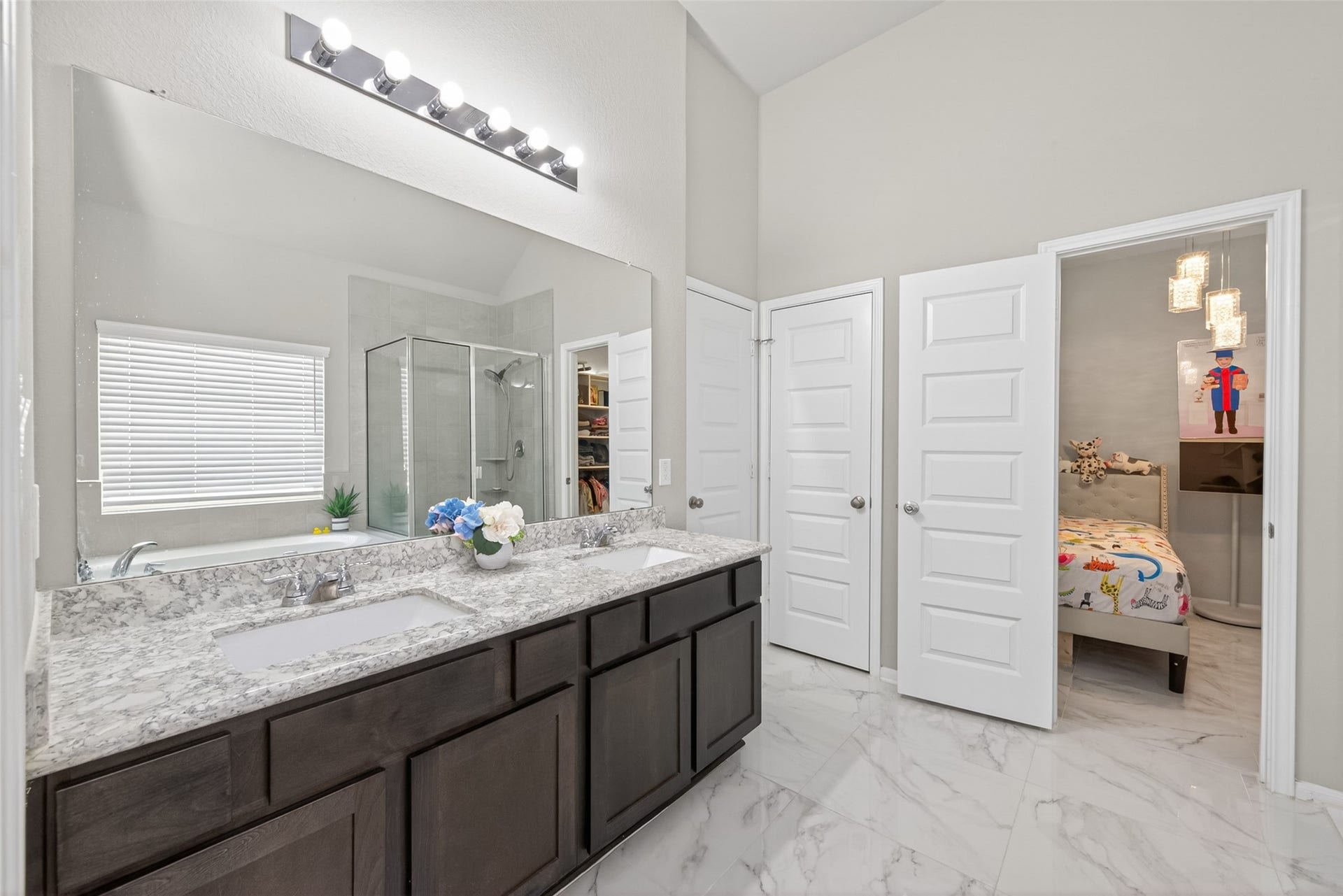 22409 Kinley Street, New Caney, TX 77357 A spacious double vanity with granite countertops, a large mirror, and a sleek glass-enclosed shower. It connects to a cozy bedroom through a white paneled door, adding convenience and style.