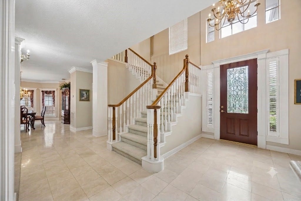Elegant foyer with a grand staircase, intricate glass door, and marble flooring, leading to a bright living and dining area.