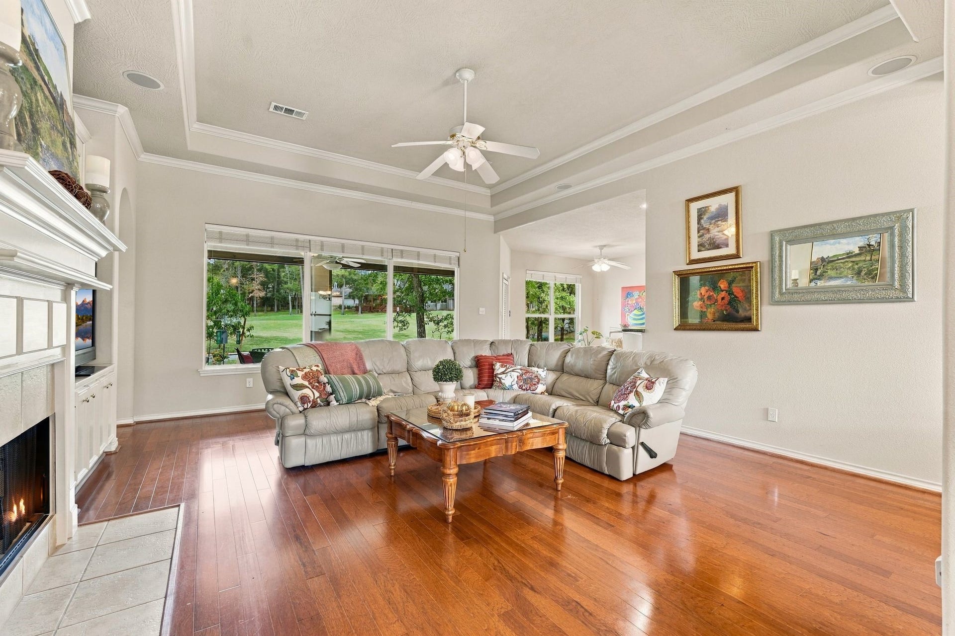 156 April Waters Drive W, Conroe, TX 77356 Spacious family room with elegant tray ceilings, featuring a cozy sectional sofa, engineered hardwood floors, and large windows offering serene views of lush greenery.