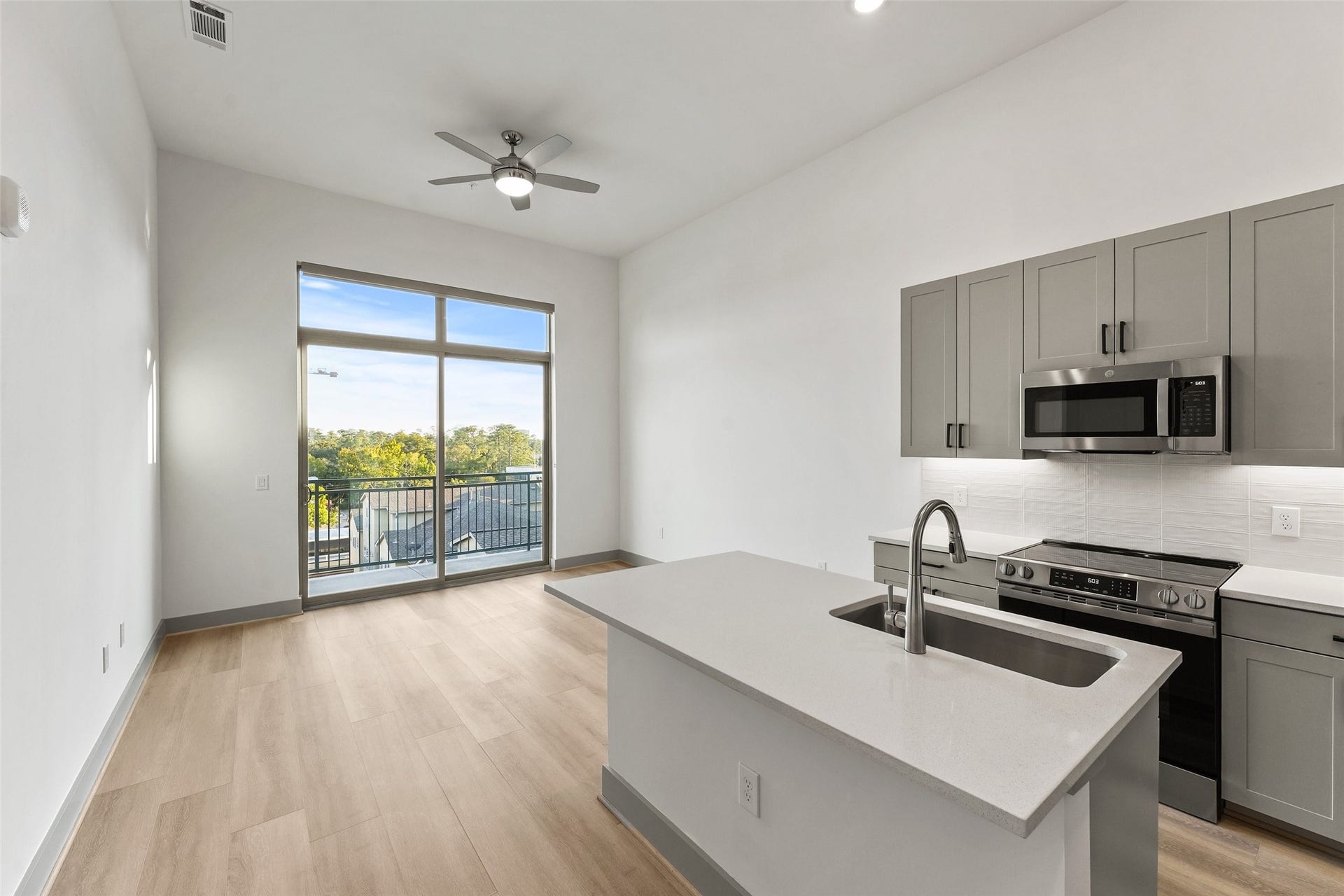170 Malone Street, #3402, Houston, TX 77007 Bright and modern open-plan kitchen and living area with sleek cabinetry, stainless steel appliances, and an island with a sink. The space features light wood flooring, a ceiling fan, and large windows leading to a balcony with a scenic view.