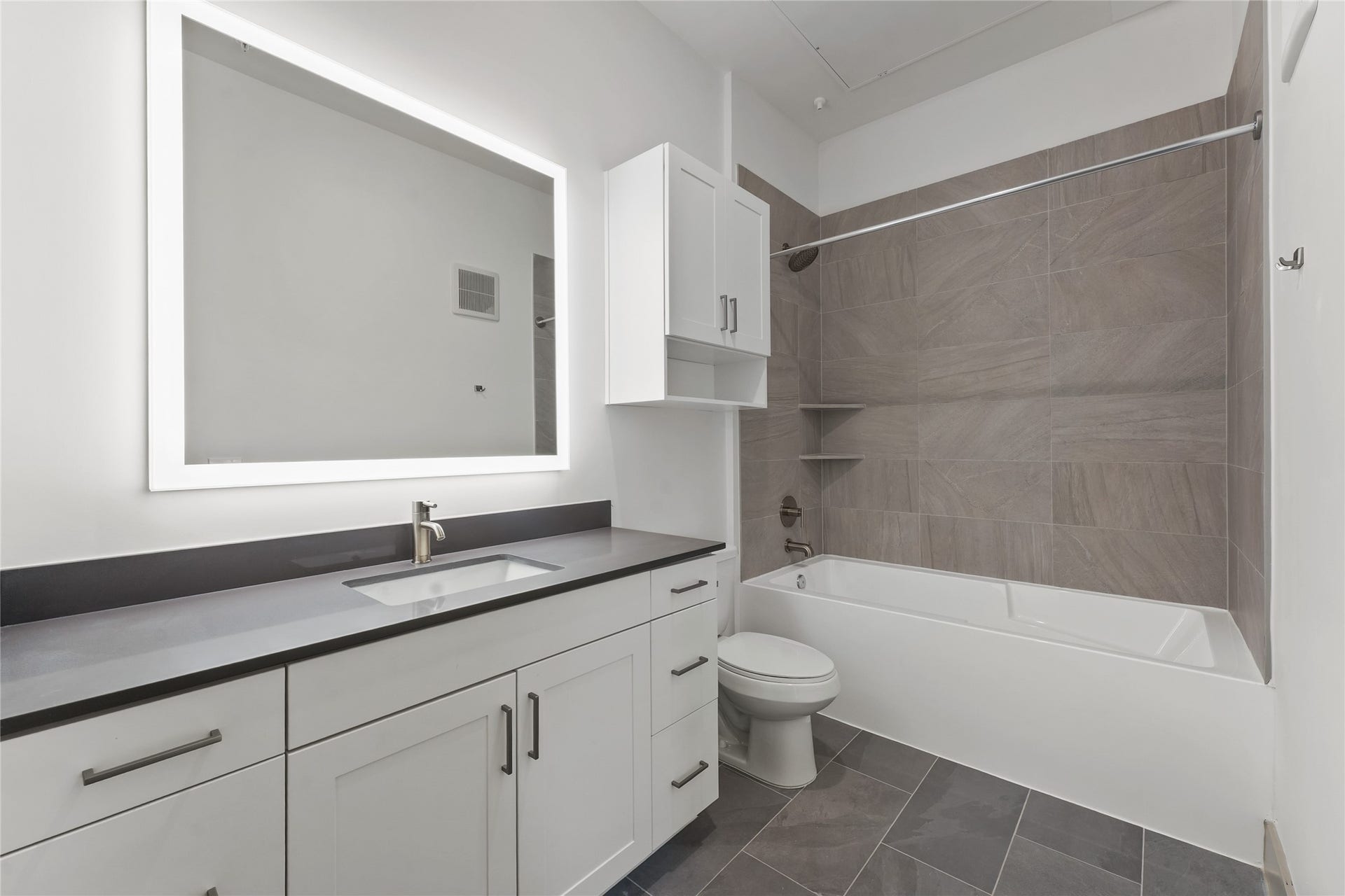 170 Malone Street, #3402, Houston, TX 77007 This modern bathroom features sleek, gray tile flooring and a spacious vanity with a large, illuminated mirror. The shower-tub combo is accented with elegant wall tiles and built-in shelving. The overall design is clean and contemporary, offering a bright and functional space.