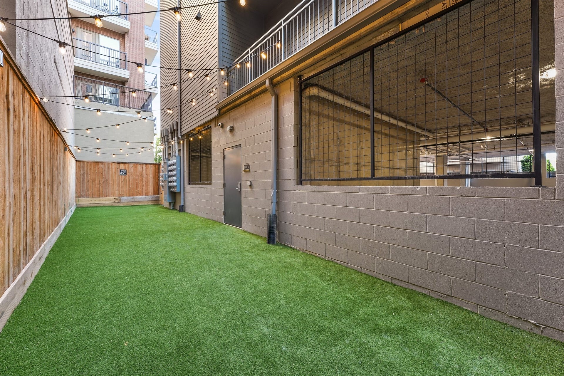 170 Malone Street, #3402, Houston, TX 77007 This photo shows a narrow outdoor space with artificial grass, enclosed by wooden fencing on one side and a concrete wall with mesh windows on the other. String lights hang above, adding a cozy ambiance, and there's a gated parking area visible through the mesh.