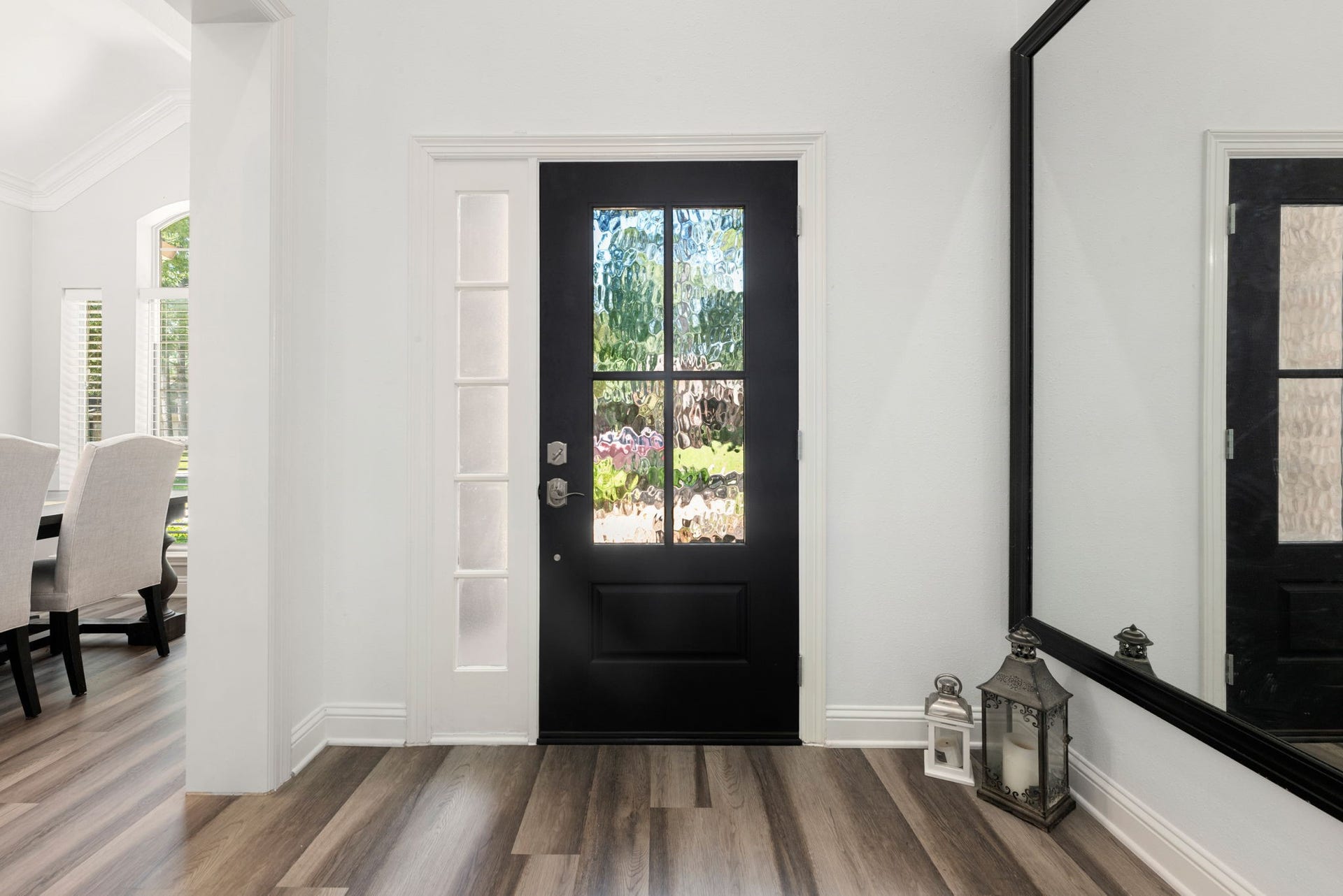 Inviting entryway featuring a striking black door with glass panels, complemented by elegant hardwood flooring , offering a glimpse into a bright dining area.