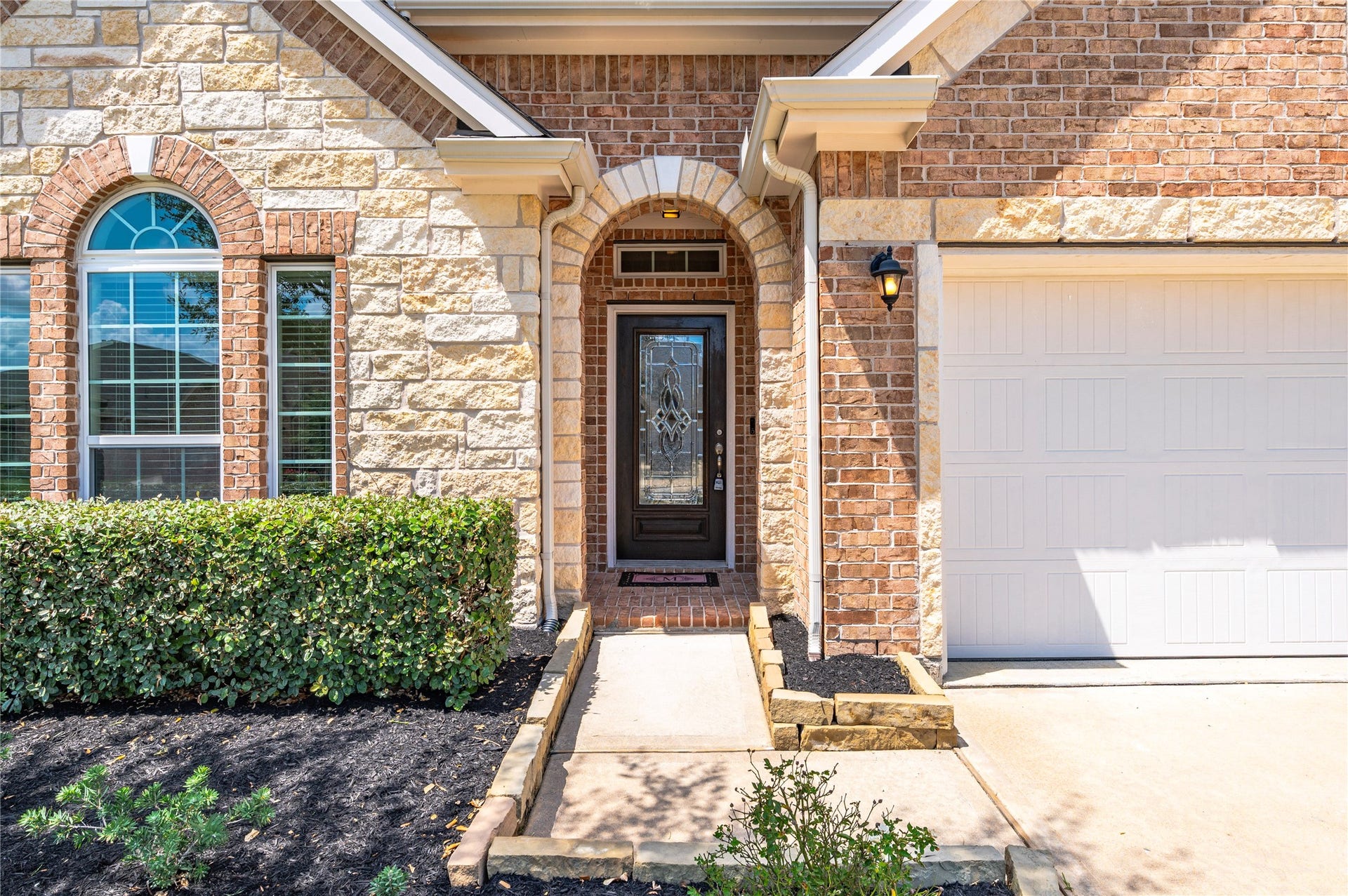 1849 Kenley Way, Alvin, TX 77511 The welcoming entrance is a blend of stone and brick, featuring an archway and elegant glass front door, complemented by manicured landscaping and a cozy porch light.