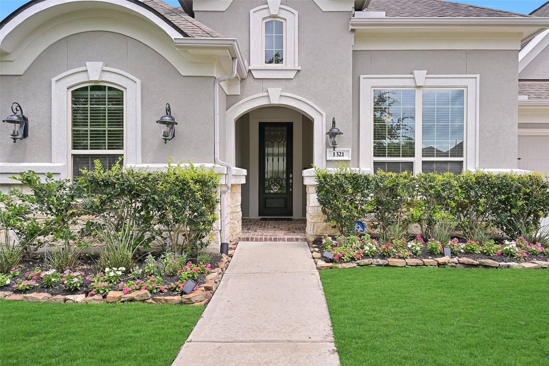 A closer look at the covered front porch highlights the elegant iron and pleated glass front door, carriage-style lantern lighting, manicured flower beds, and architectural detailing that sets the tone for the quality found throughout the home.