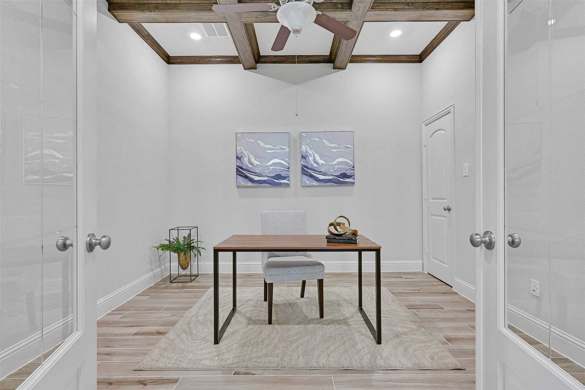 Perfect for working from home, this study features wood-look tile flooring, a coffered ceiling with rich beam detail, recessed lighting, a ceiling fan, French doors with glass inserts, and a convenient closet for additional storage.