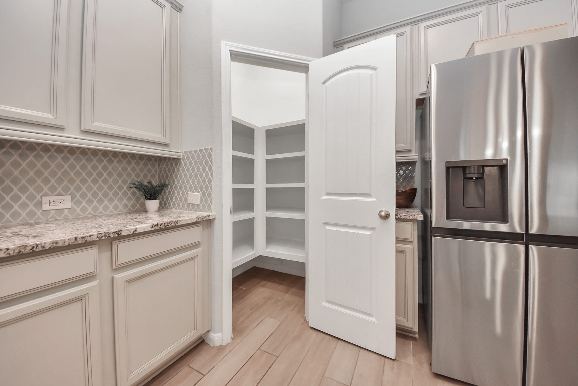 This photo showcases a modern kitchen with light-colored cabinets, a sleek stainless steel refrigerator, and a spacious walk-in pantry with ample shelving. The countertops are granite, and the backsplash features a stylish tile pattern, enhancing the overall aesthetic.