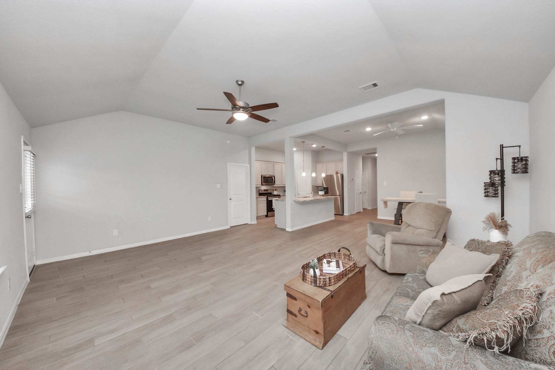 This spacious open-plan living area features light wood flooring and neutral walls, creating a bright and airy feel. The room includes a ceiling fan, cozy seating, and a view into the modern kitchen, which boasts stainless steel appliances and a breakfast bar.