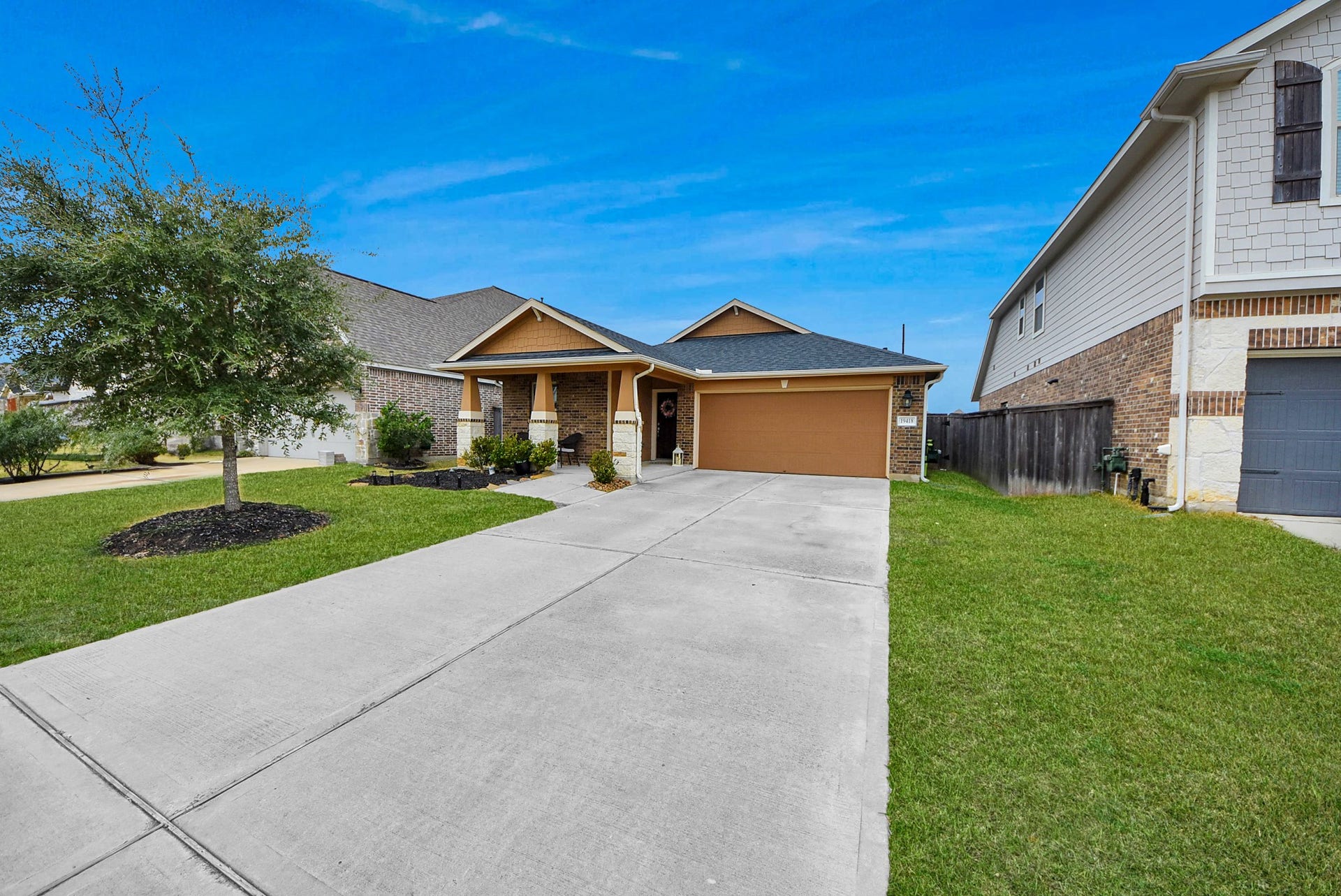 This is a charming single-story home with a brick facade and a gabled roof. It features a two-car garage and a spacious driveway. The front yard is neatly landscaped with a small tree and shrubs.