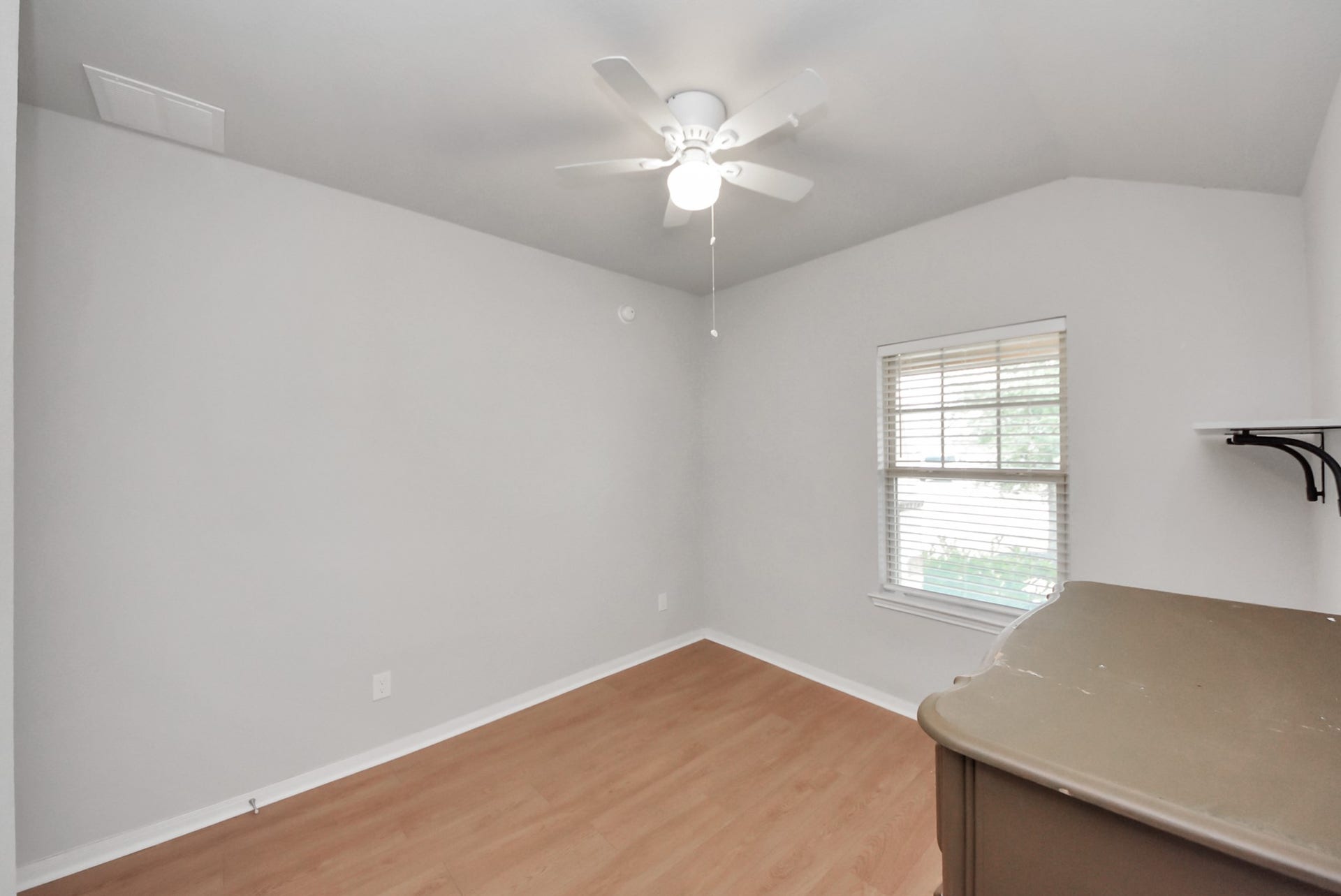This photo shows a small, bright room with light-colored walls and wood flooring. It features a ceiling fan and a window with blinds, offering natural light. A small shelf is mounted on the wall.