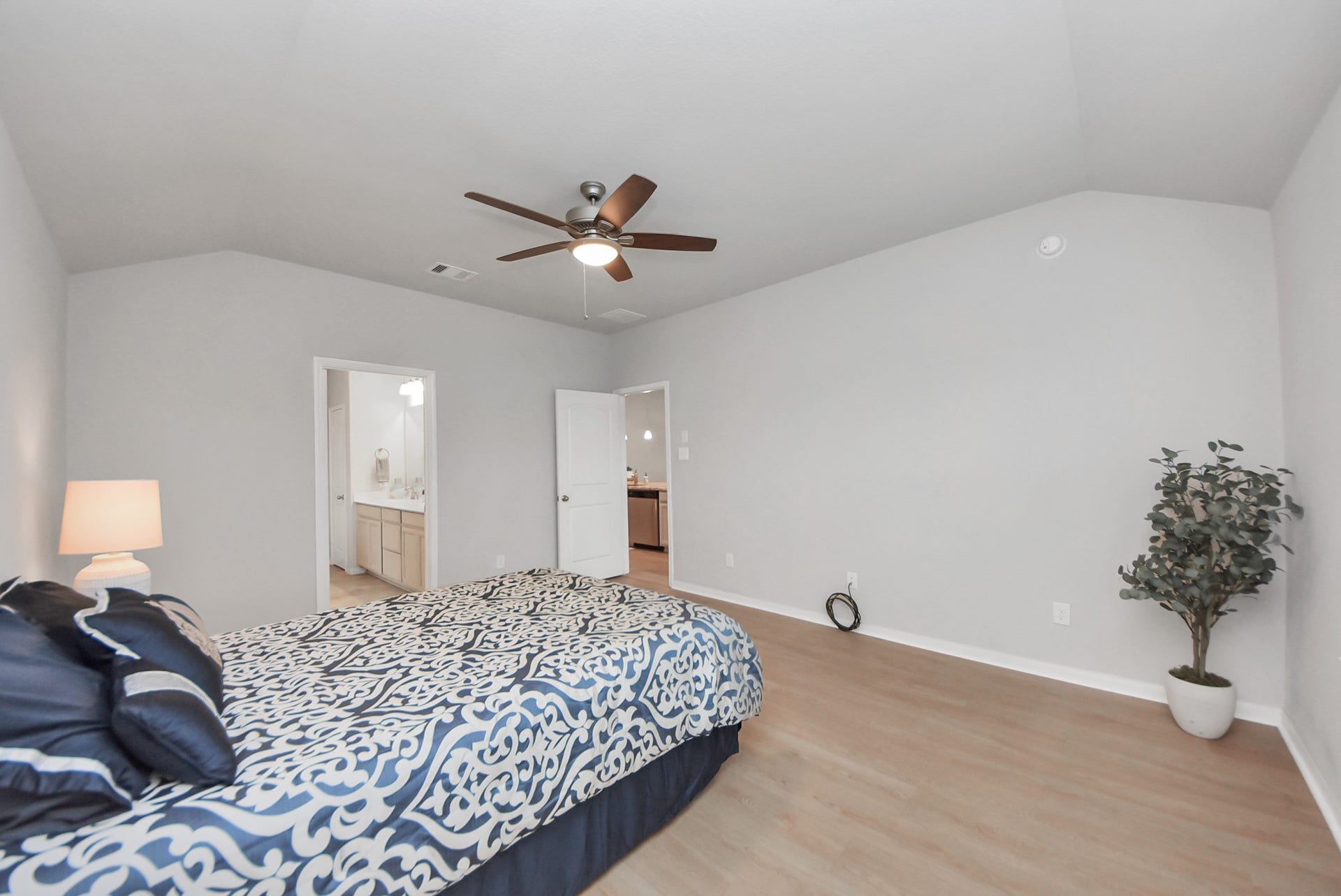 This bedroom features a modern ceiling fan, neutral walls, and wood flooring. It includes a cozy bed, a bedside lamp, and an en-suite bathroom. A potted plant adds a touch of greenery.