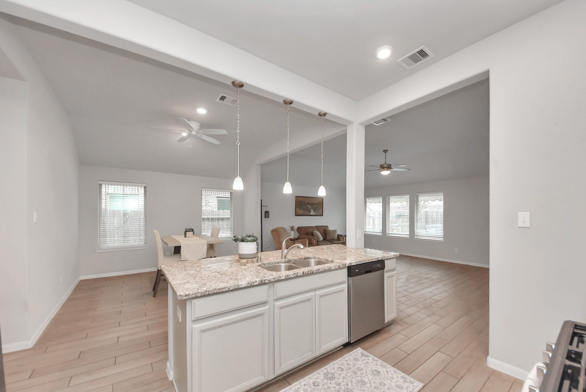 This photo showcases a spacious open-concept living area with a modern kitchen. It features a large island with granite countertops, pendant lighting, and stainless steel appliances. The adjoining living room and dining area have ample natural light, wood-like flooring, and ceiling fans, creating a welcoming atmosphere.