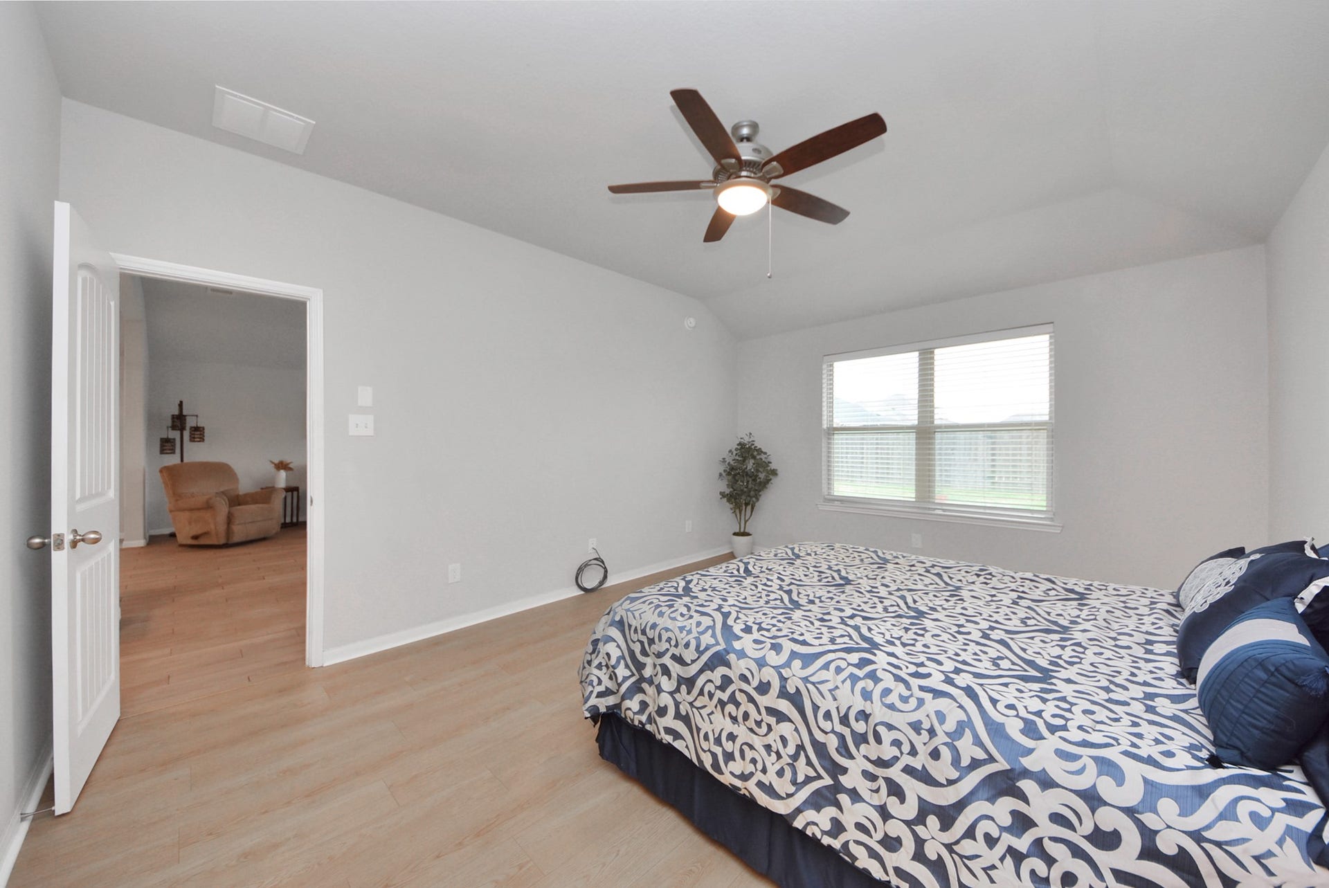 This spacious Primary bedroom features a vaulted ceiling with a central fan, ample natural light from a large window, and light wood flooring. It's furnished with a cozy bed and offers an open doorway leading to a comfortable sitting area.