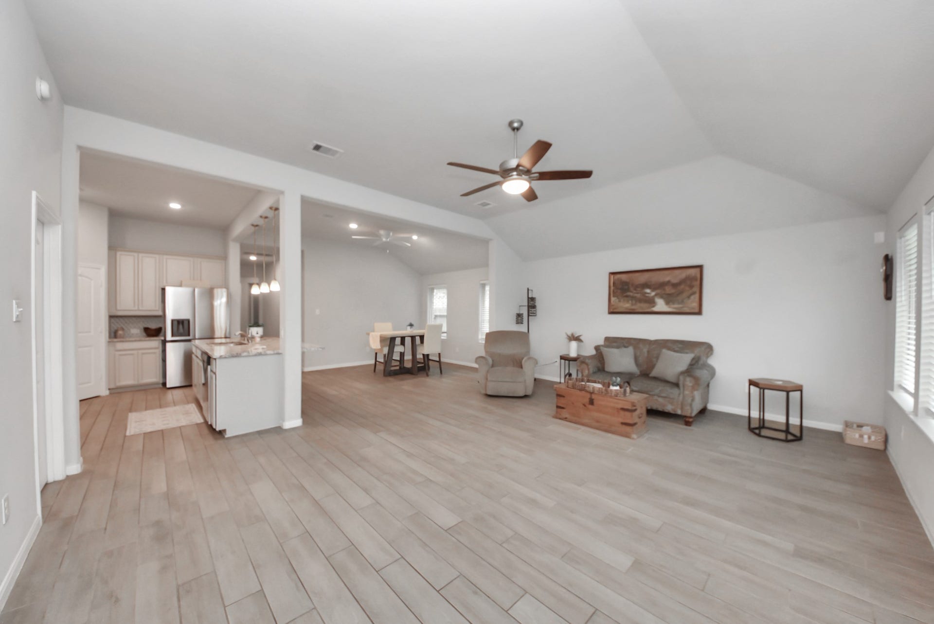 This spacious open-concept living area features light wood flooring, a modern kitchen with stainless steel appliances, and ample natural light from large windows. The room includes a cozy seating area, ceiling fan, and neutral decor, ideal for comfortable living and entertaining.