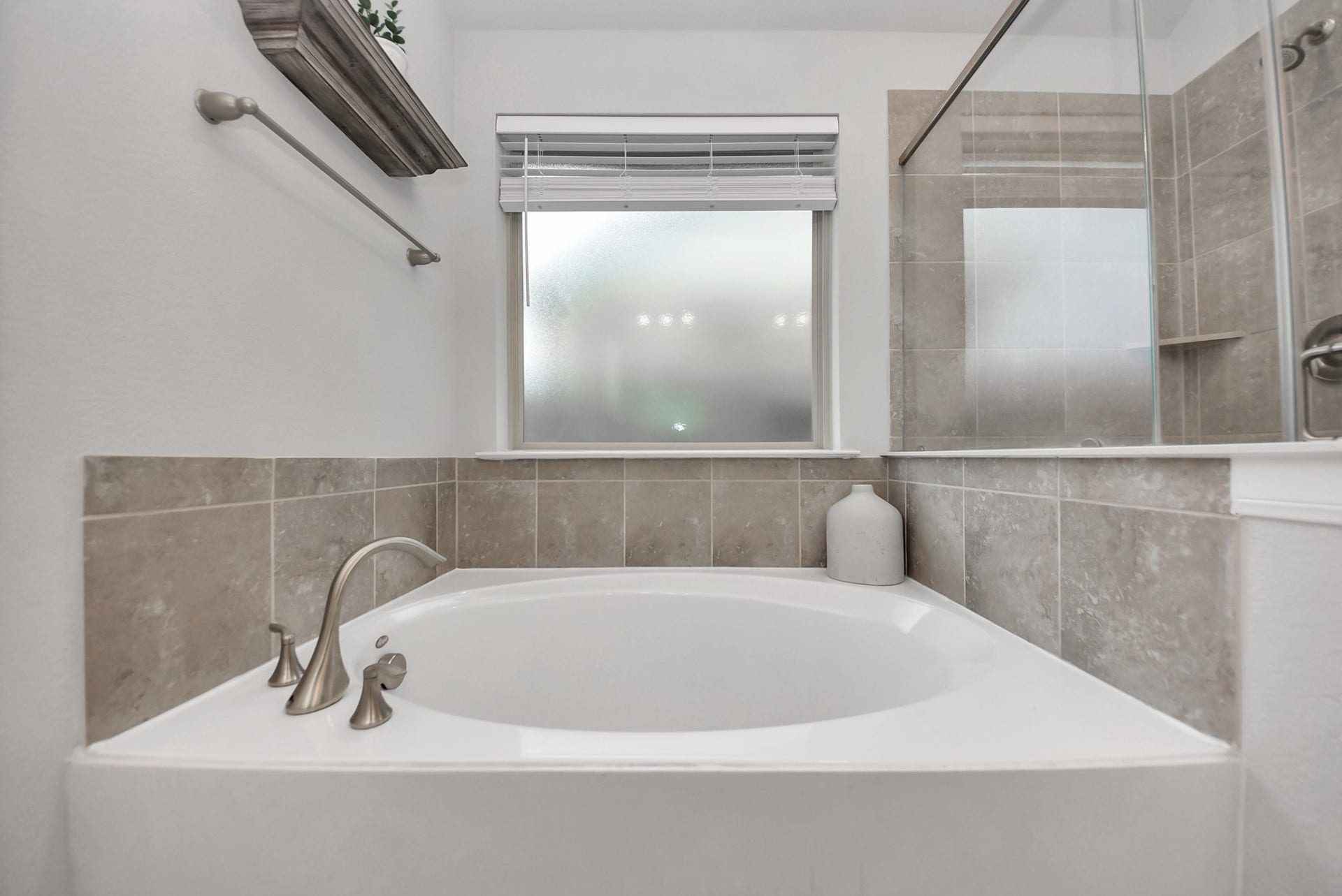 This Owners bath features a modern design with a spacious white bathtub, sleek brushed nickel fixtures, and elegant beige tile accents. A frosted window provides privacy and natural light, complemented by a glass shower enclosure for a contemporary feel.