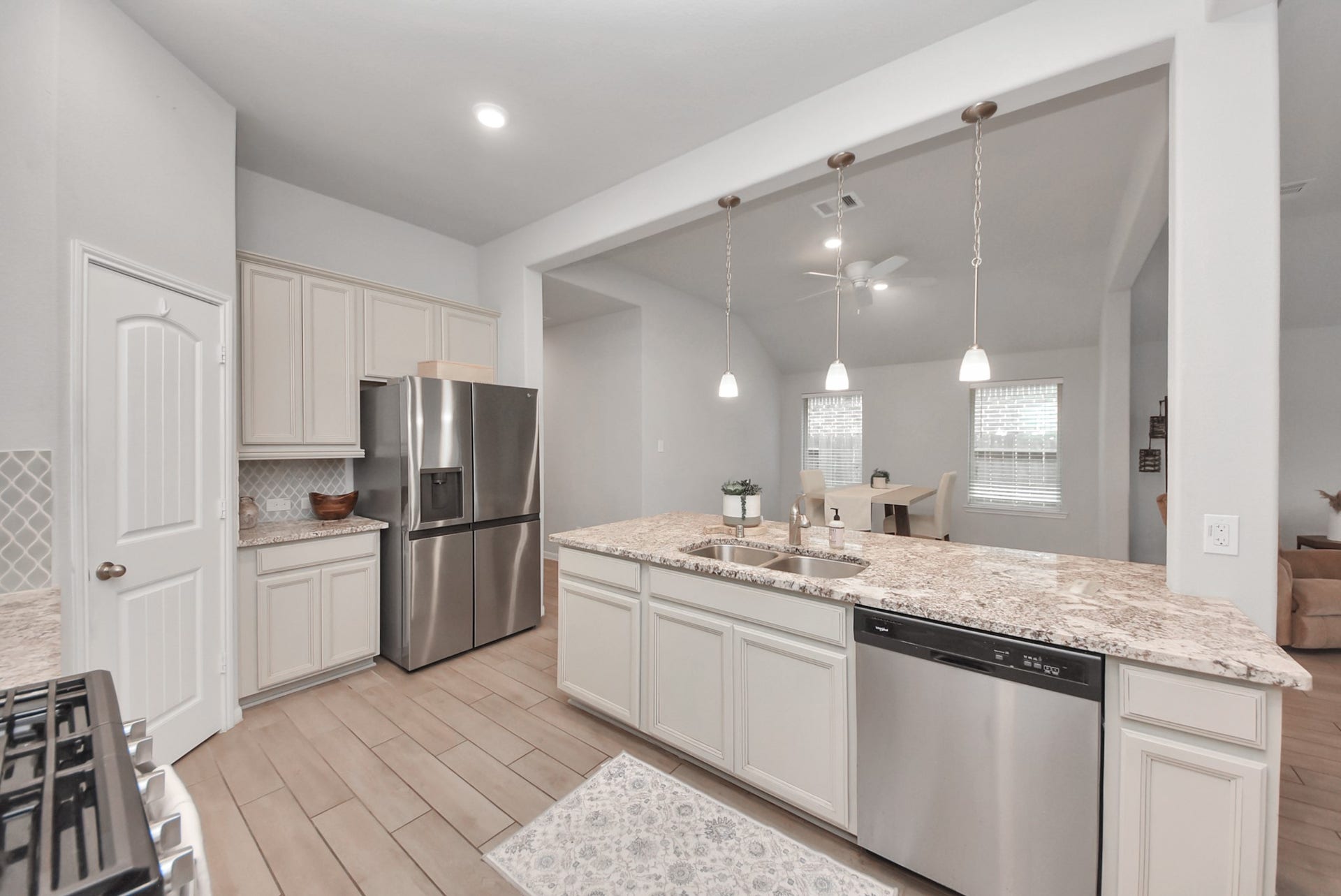 This modern kitchen features stainless steel appliances, a spacious granite island with a sink, and pendant lighting. It opens up to a bright dining area with large windows.