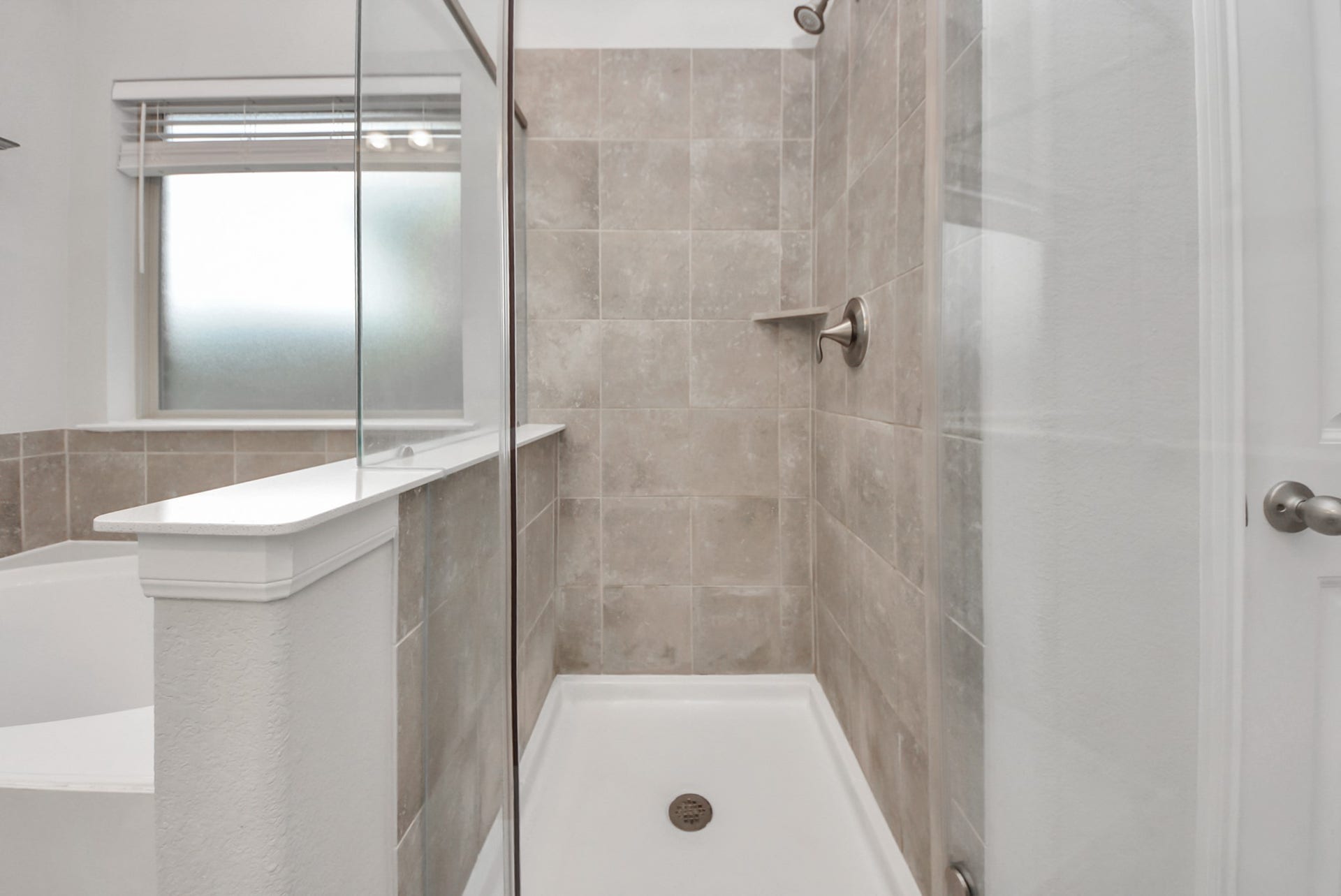 This photo shows a modern bathroom with a glass-enclosed shower featuring neutral-toned tiles, a built-in shelf, and a frosted window for privacy.