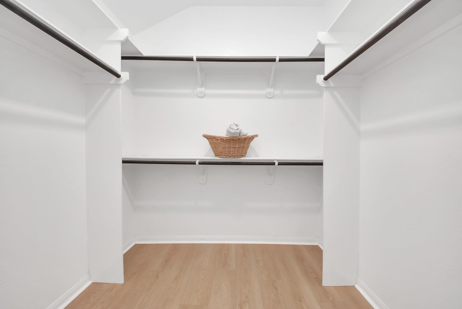 This photo shows a spacious walk-in closet with white walls and wooden flooring. It features multiple hanging rods for clothing and a shelf with a decorative basket, offering ample storage space.