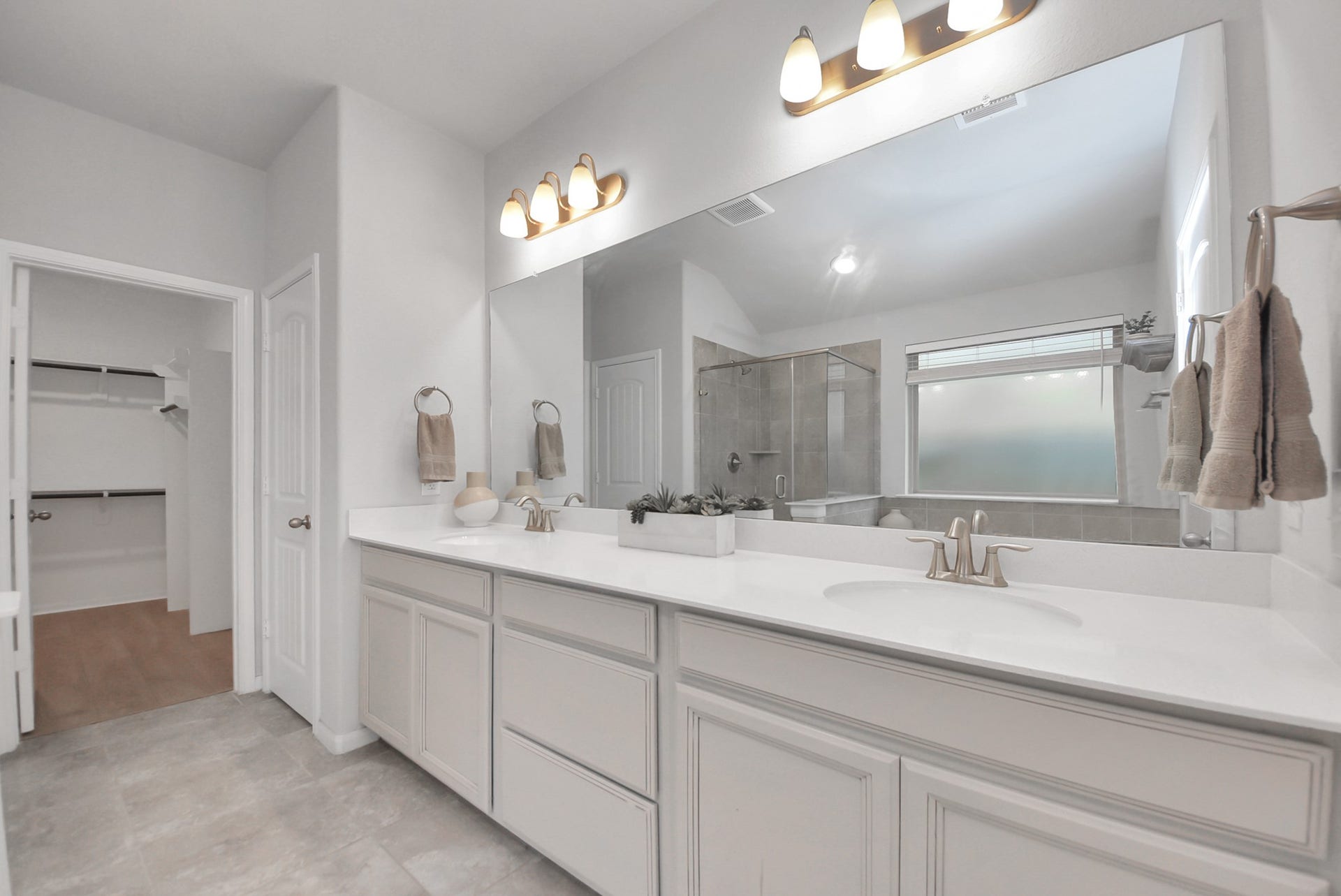 This spacious modern bathroom features double sinks with ample counter space, elegant lighting, a large mirror, and a walk-in shower. It also includes a convenient entry to a walk-in closet, offering plenty of storage.
