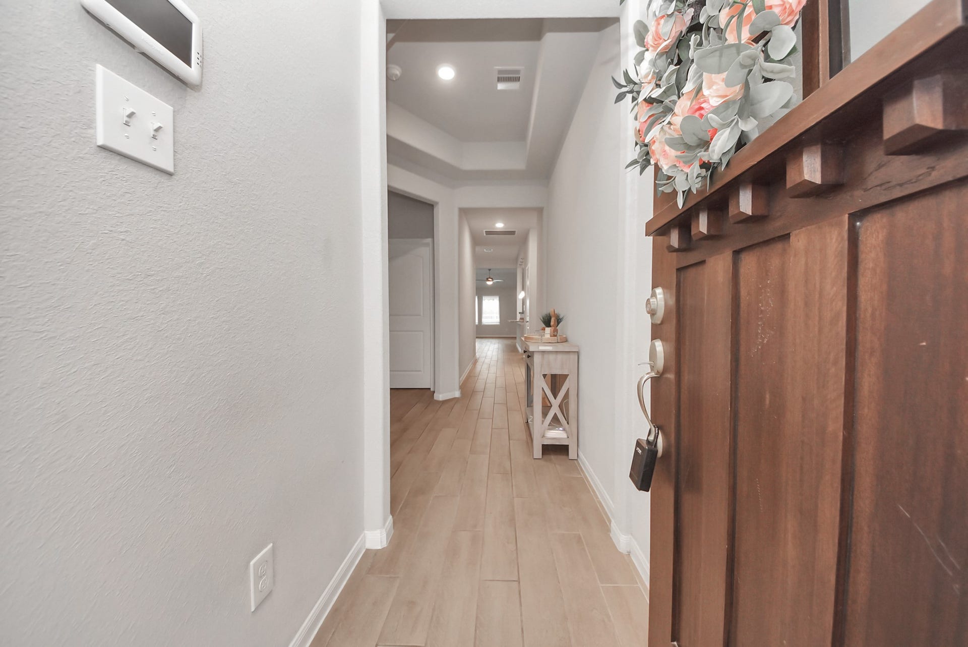 This photo showcases a welcoming entrance hallway with light wood flooring, neutral walls, and recessed lighting. A wooden front door with a floral wreath opens into the space, which features a decorative console table. The hallway leads to a bright room at the end, creating an inviting first impression.