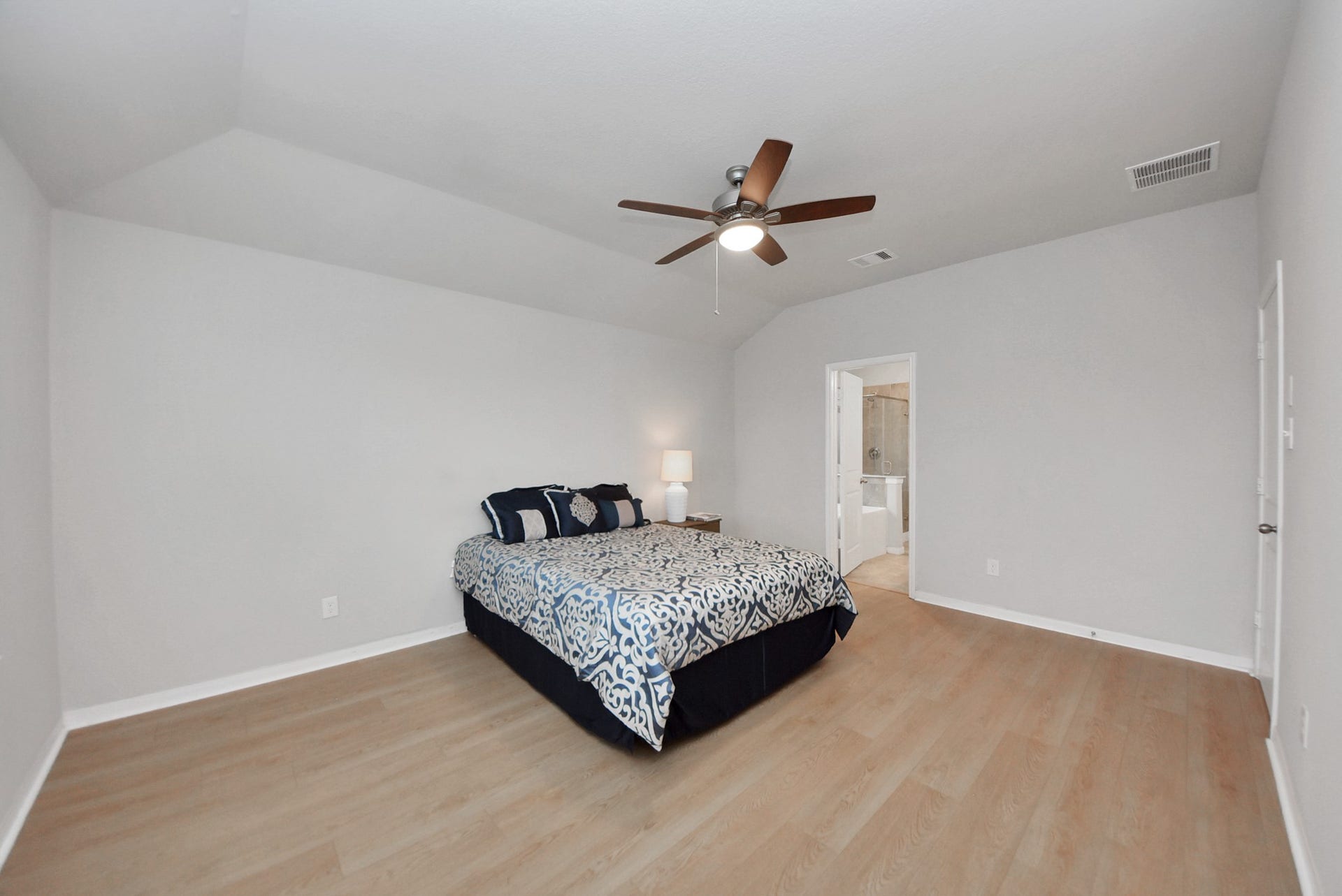 This photo captures spacious bedroom with light wood flooring, neutral walls, and a ceiling fan. The room features a neatly made bed and an en suite bathroom, offering a clean and modern look.