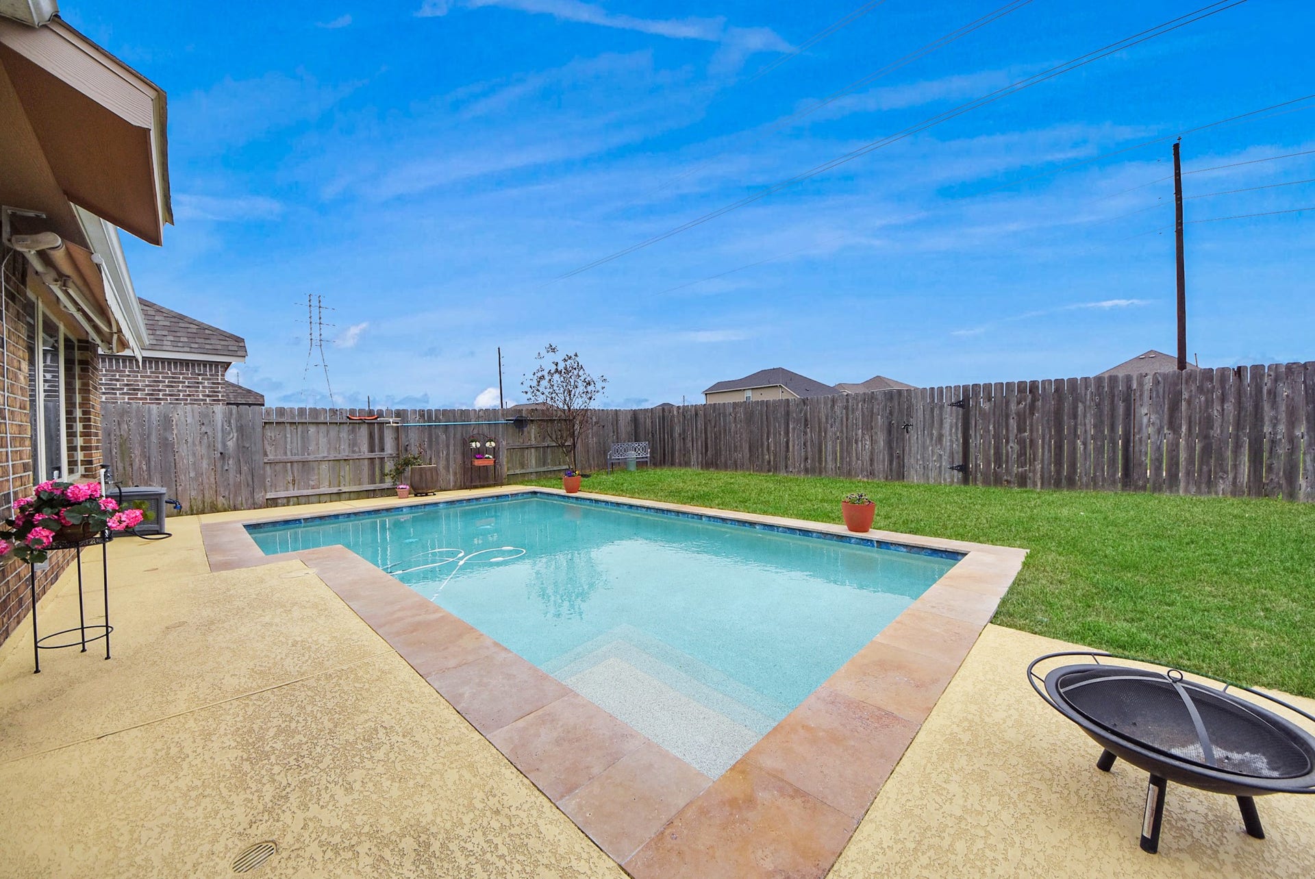 This backyard features a spacious in-ground pool surrounded by a clean concrete deck and a well-maintained lawn. The area is enclosed with a wooden fence for privacy, and there's a small seating area, perfect for relaxation or entertaining.