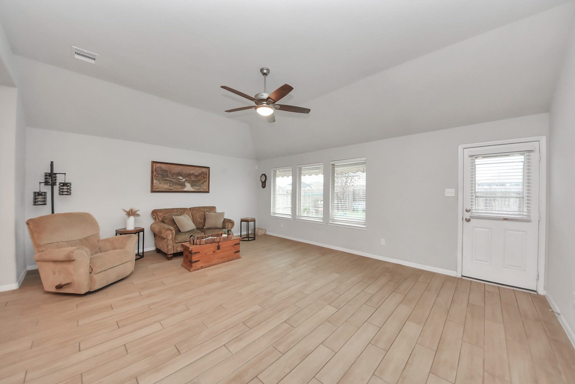 This spacious living room features light wood flooring, neutral walls, and ample natural light from large windows. It includes a cozy seating area with a sofa and armchair, a ceiling fan for comfort, and access to the outdoor space through a door on the right.